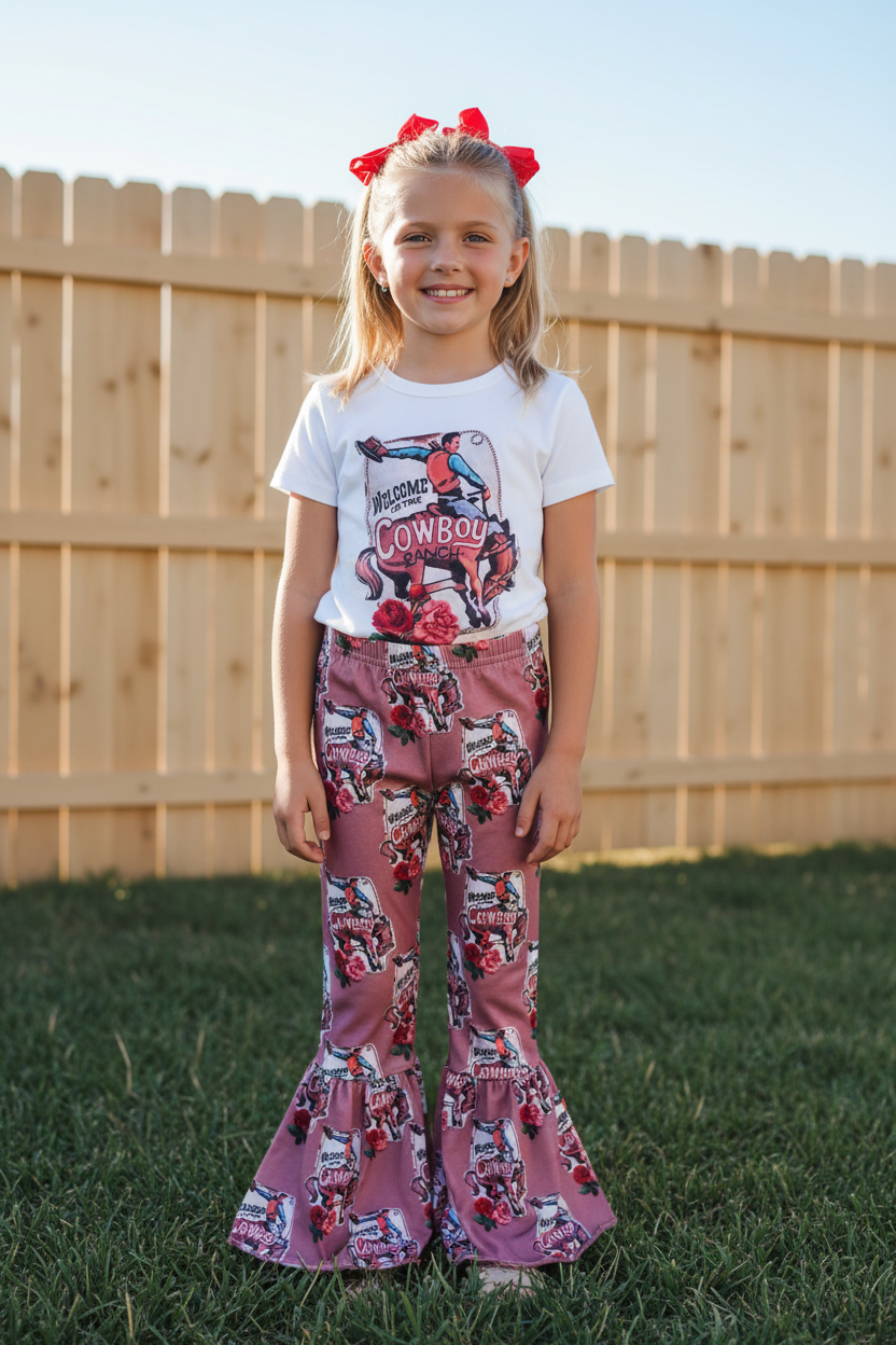 Young girl wearing a white t-shirt with graphics and pink flared pants standing in front of a wooden fence.