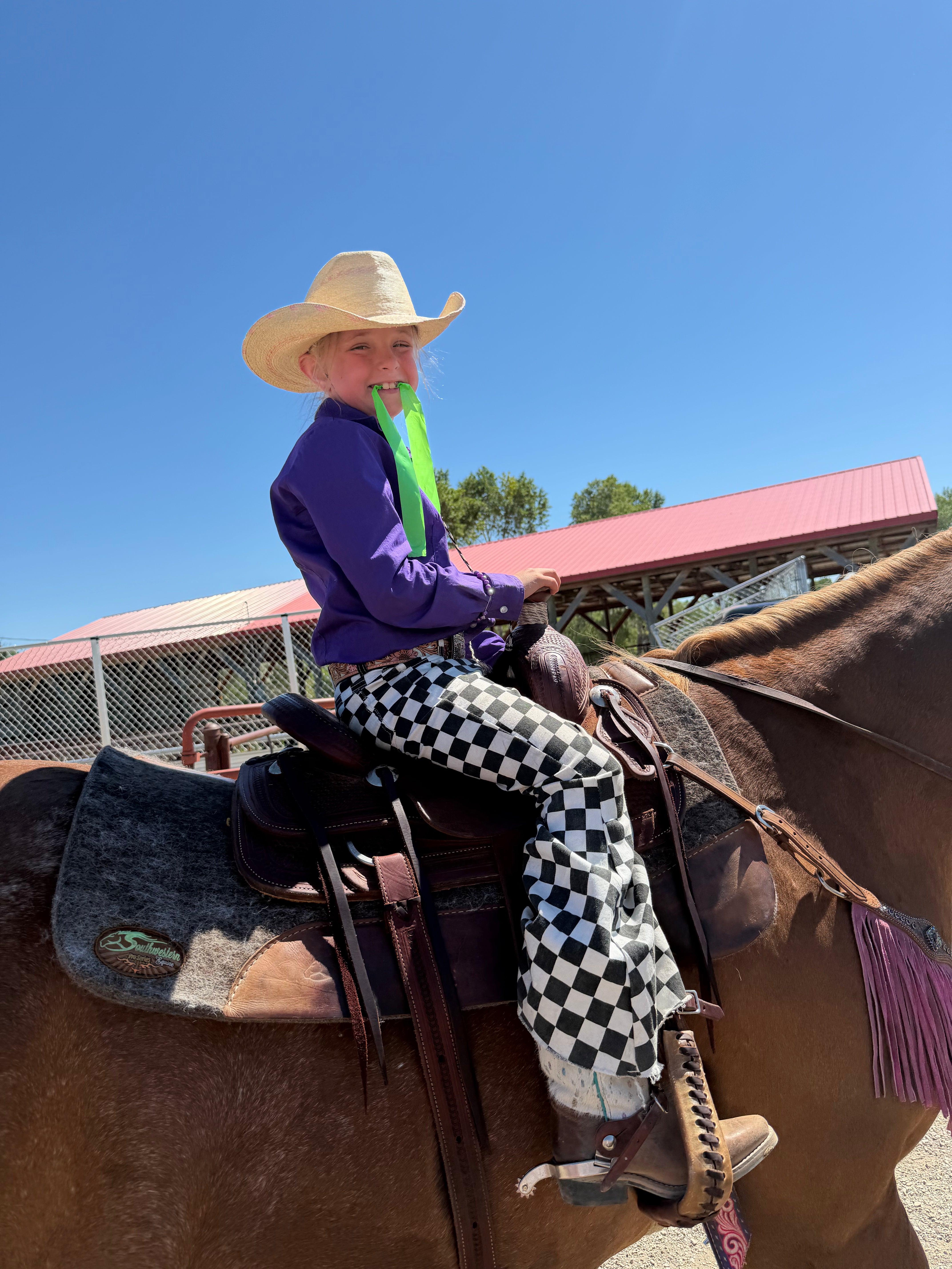 A young girl wearing a straw cowboy hat, purple long-sleeve shirt, neon green neck gaiter, and black-and-white checkered pants sits on a brown horse outdoors under a clear blue sky.