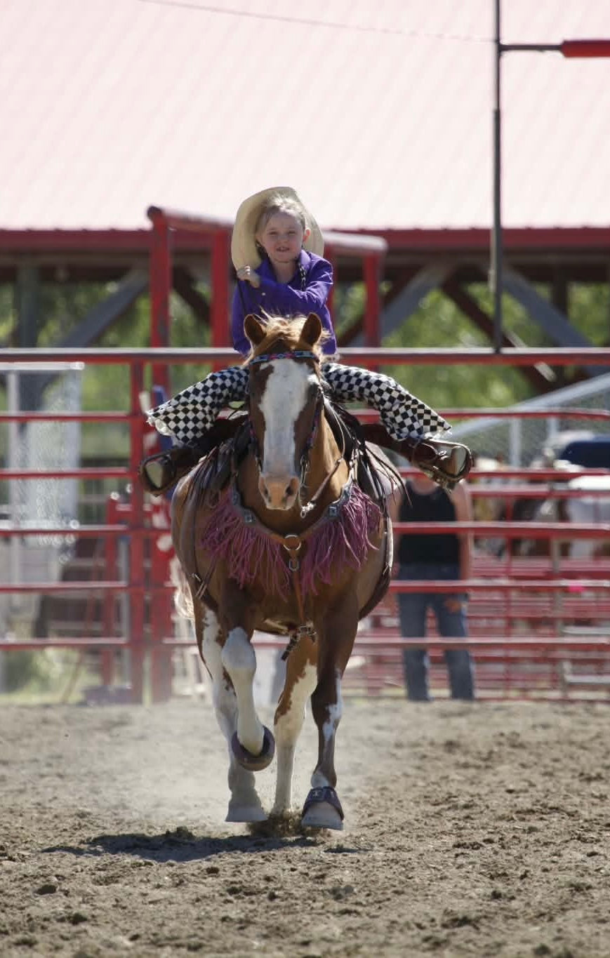 A young girl wearing a cowboy hat, purple shirt, and black-and-white checkered pants rides a brown and white horse in a dirt arena at an rodeo, with red metal fences and a structure in the background.
