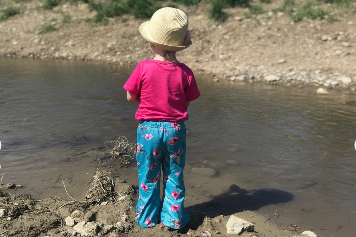 GIRLS WEARING HOT PINK TSHIRT AND TURQUOISE COWSKUL FLOWER BELL BOTTOM PANTS STANDING AT CREEK. Available in toddler sizes 2T–5T, these boho bell bottoms are made for spring and summer play, photos, and everyday wear.