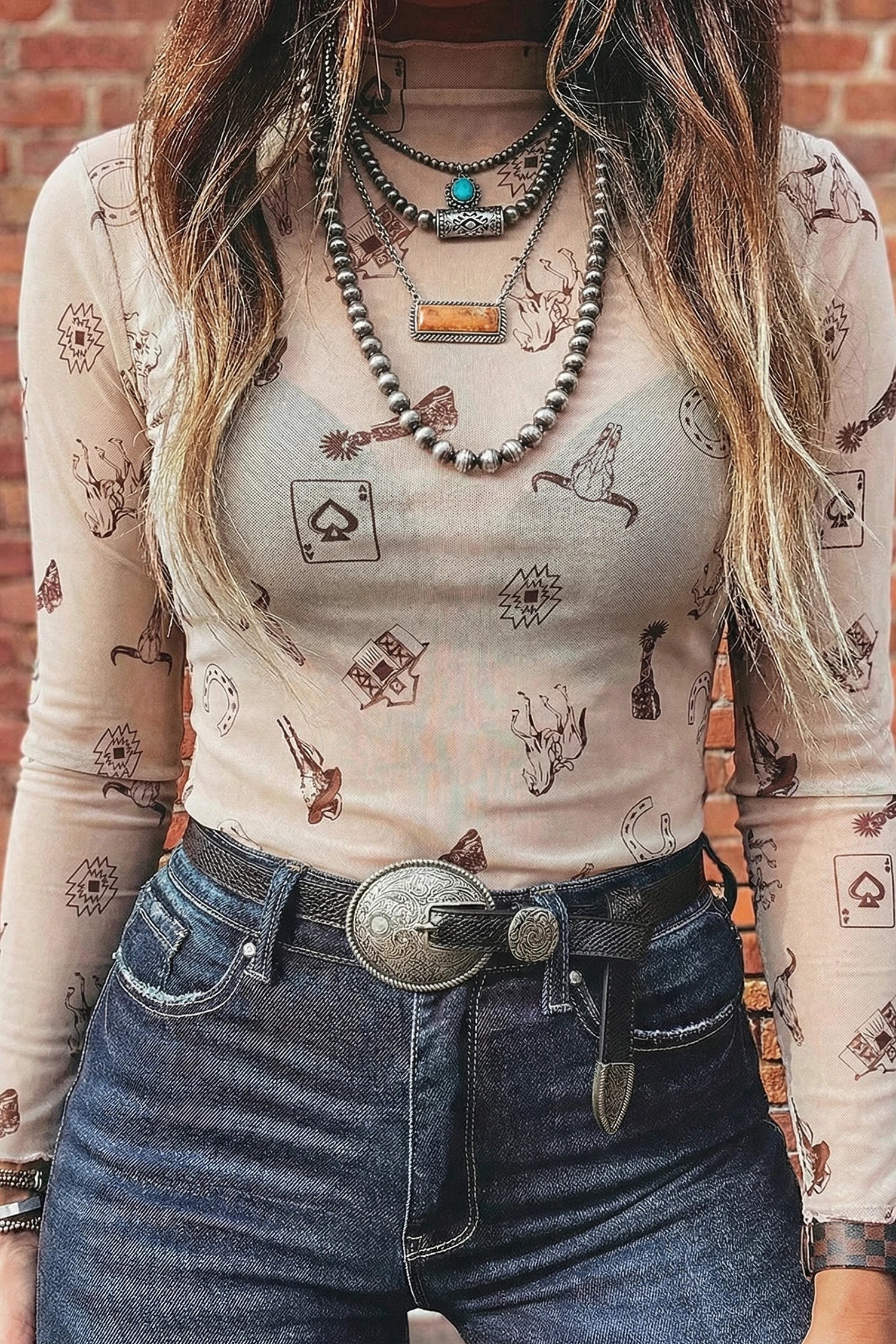WOMAN wearing a patterned long-sleeve MESH top FEATURING HORSES, AZTEC AND COW SKUL jeans with a belt, standing against a brick wall.