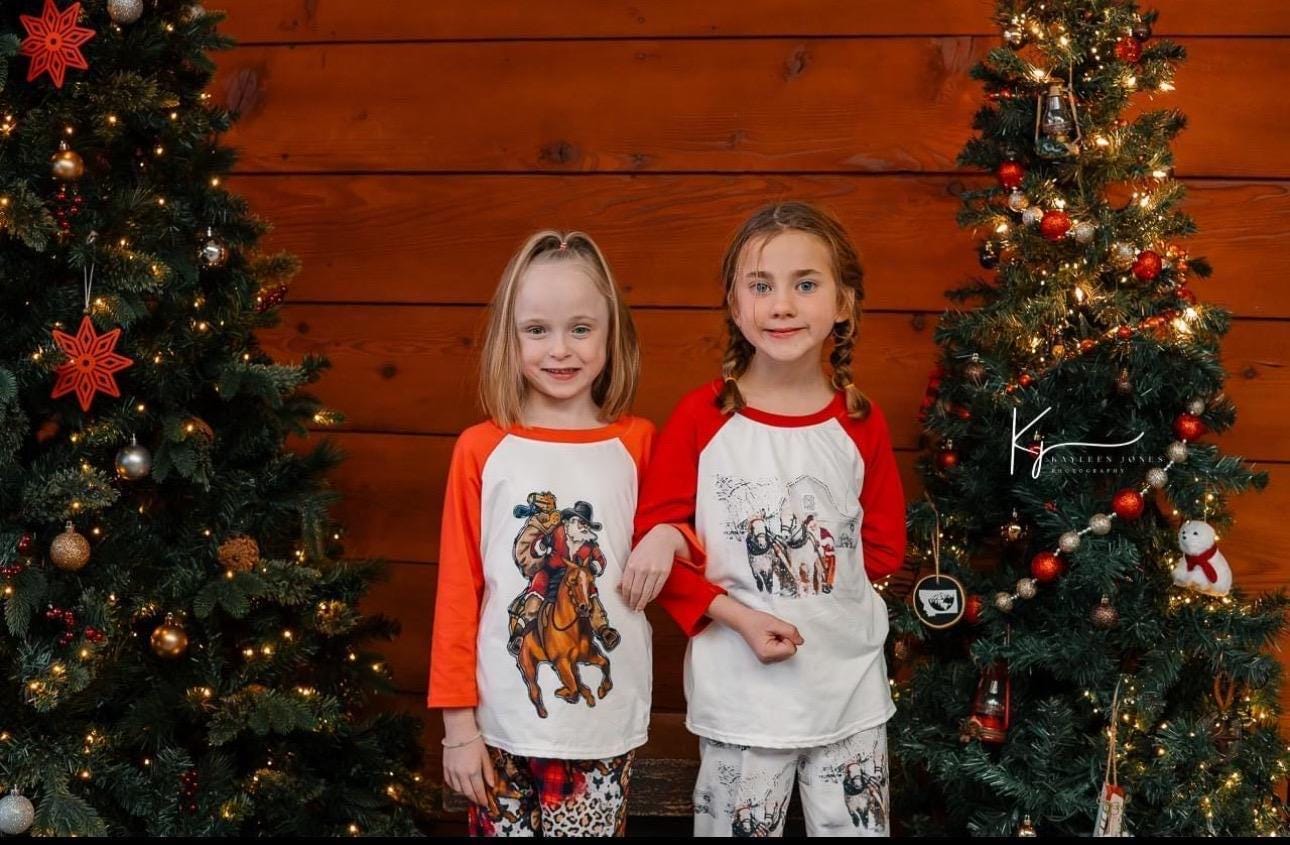 2 girls modeling baseball sleeve style cowboy santa pajama tops in fromt of decorated christmas trees