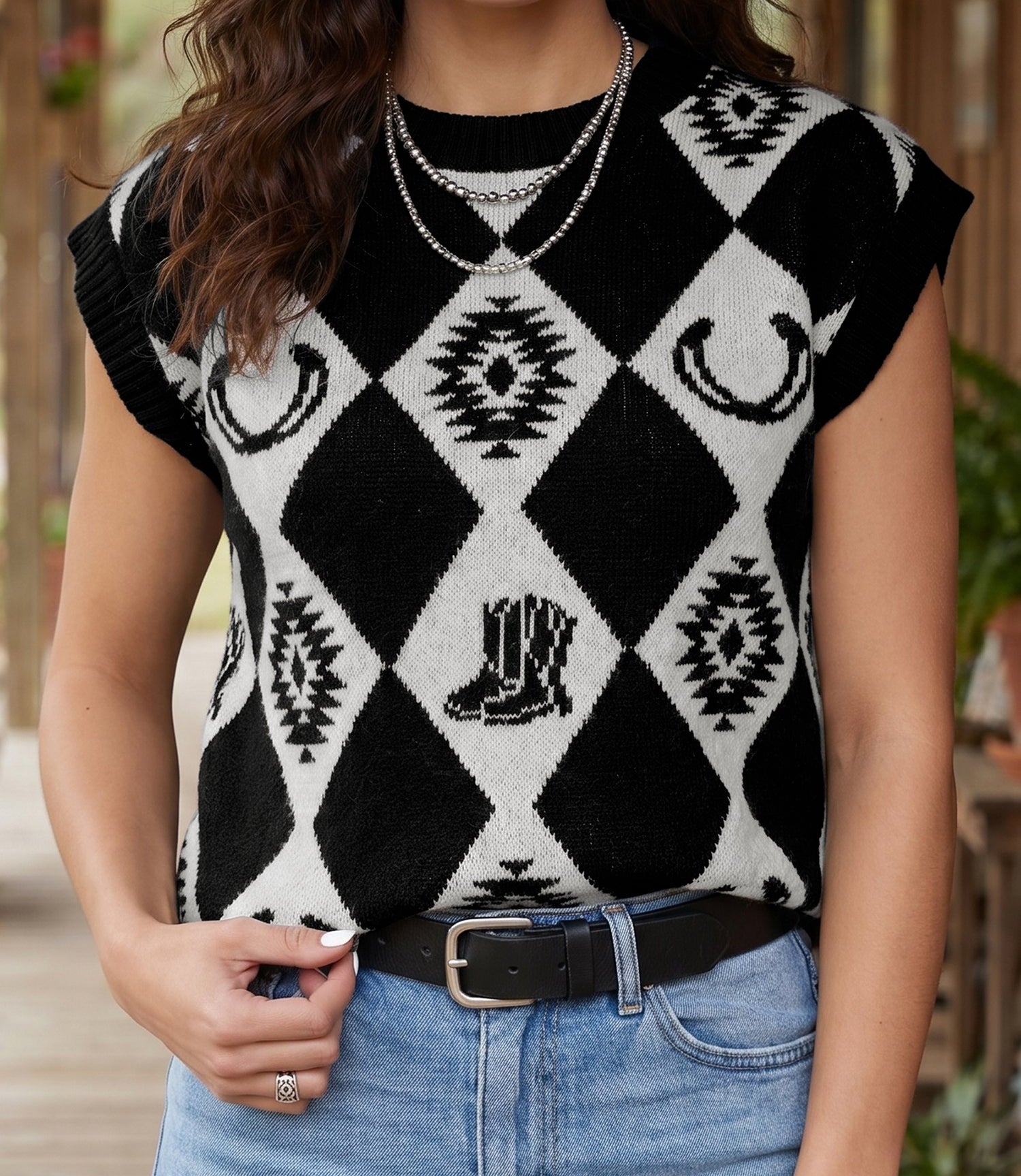 A woman wearing a black and white argyle knit sweater with western-themed motifs including horseshoes, boots, and geometric patterns, paired with light blue jeans and layered necklaces.