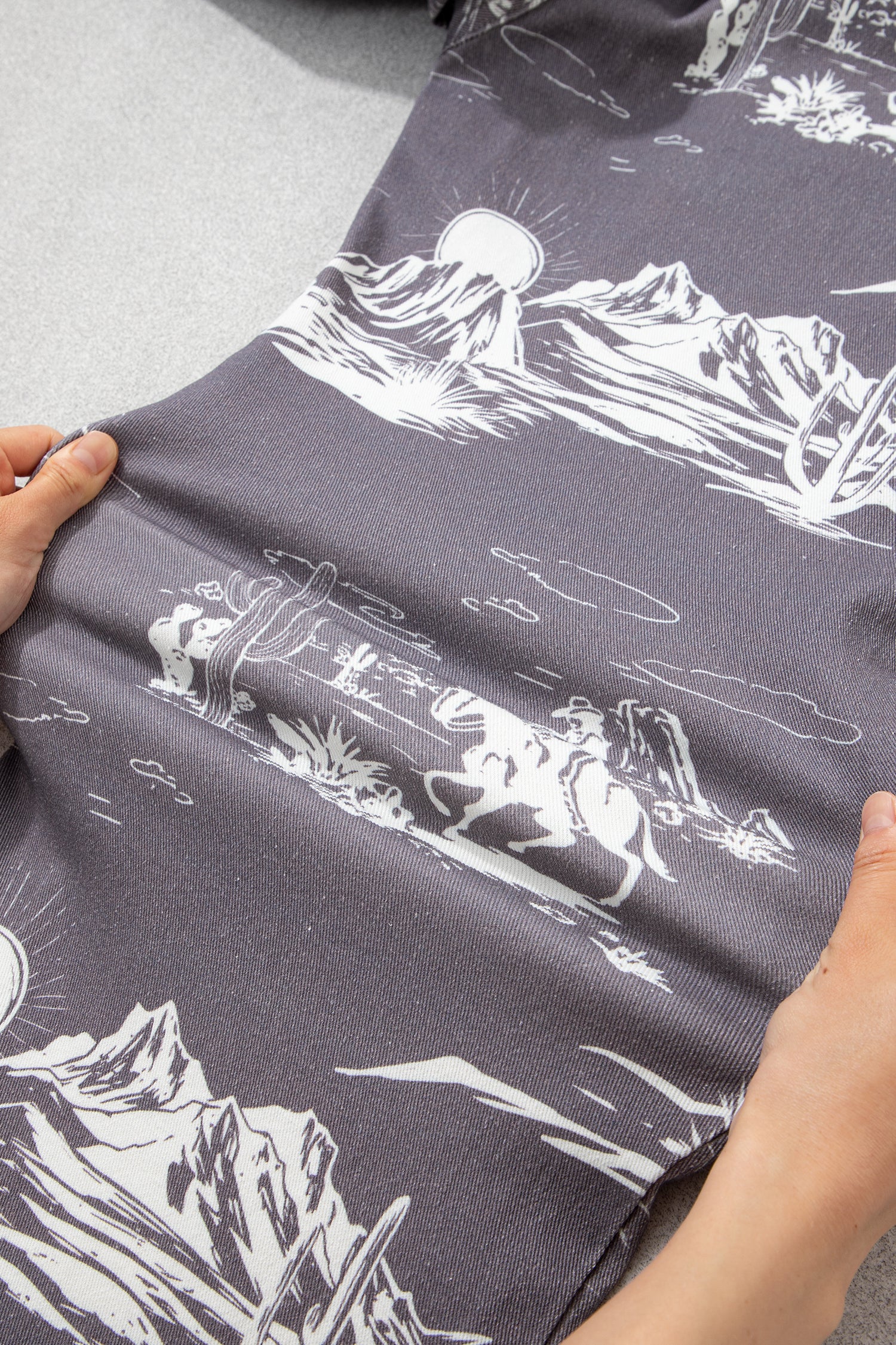 Person holding a dark fabric with white mountain and landscape designs showing how stretchy the fabric is by pulling the pant leg