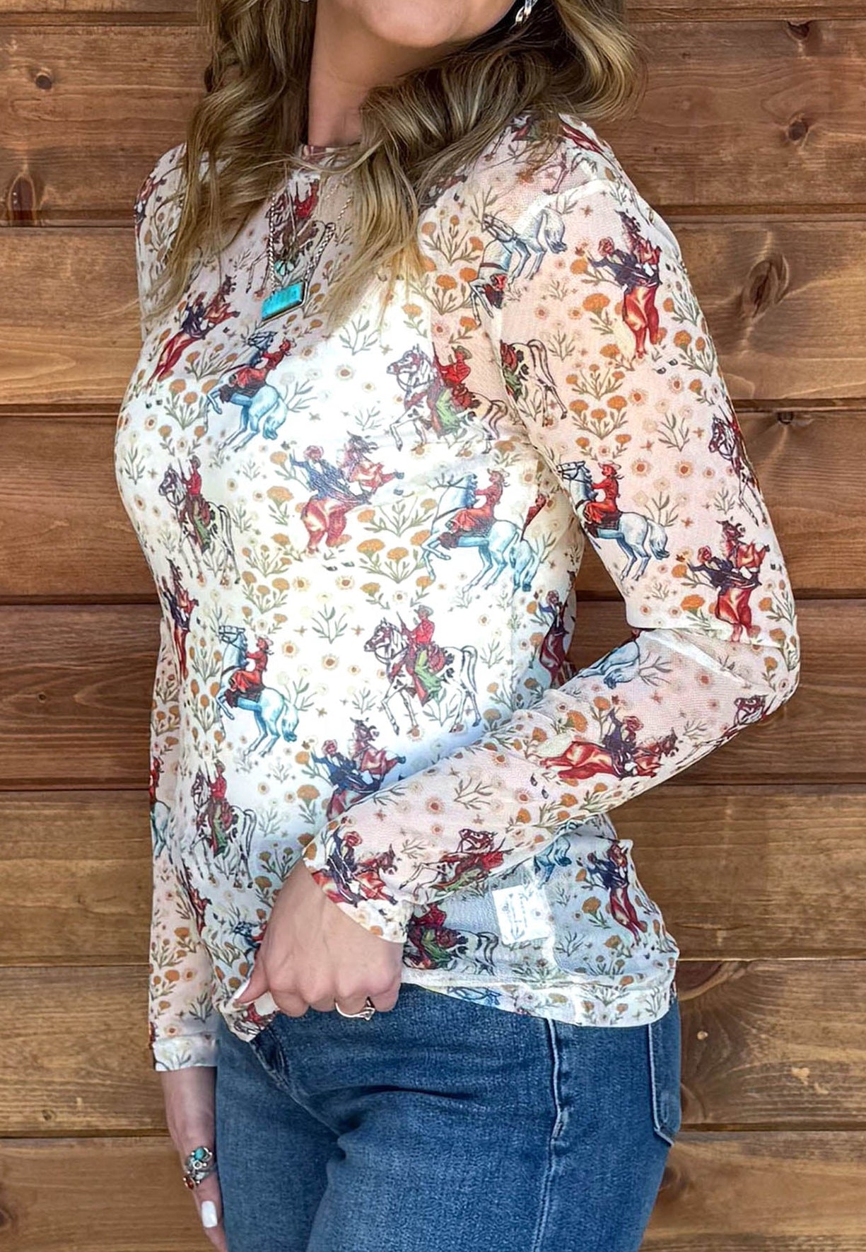 A woman wearing a long-sleeve floral and cowboy print top with blue jeans, standing against a rustic wooden plank wall.