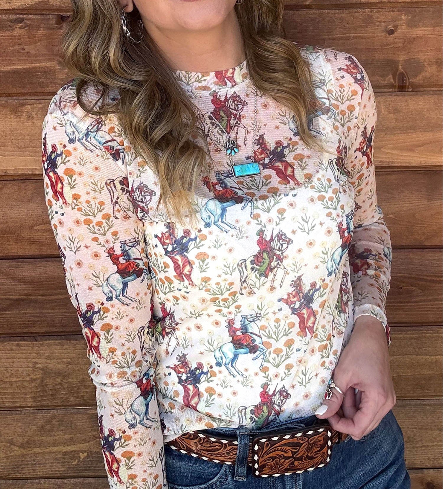 A woman with blonde hair stands against a rustic wooden plank wall wearing a long-sleeve western-style blouse with a multicolor cowboy and cattle print on a cream background, paired with blue jeans and a brown concho belt.