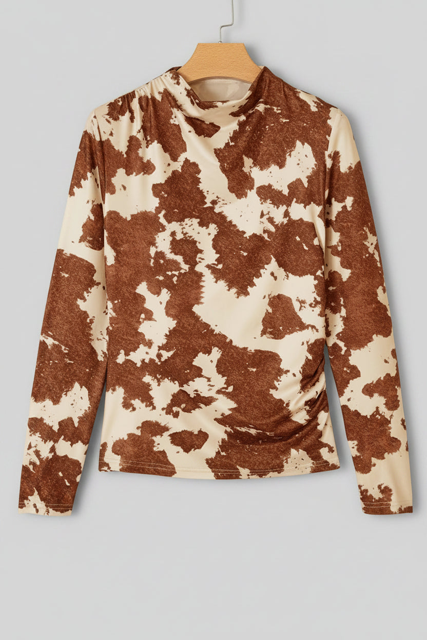 A long-sleeve mock neck top with a brown and cream cow print pattern, hanging on a wooden hanger against a plain off-white wall.