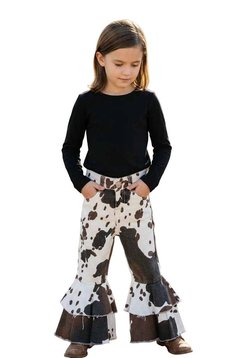 A young girl modeling a pair of cow-print flared pants with tiered ruffle hems, paired with a black long-sleeve top, against a solid white background.