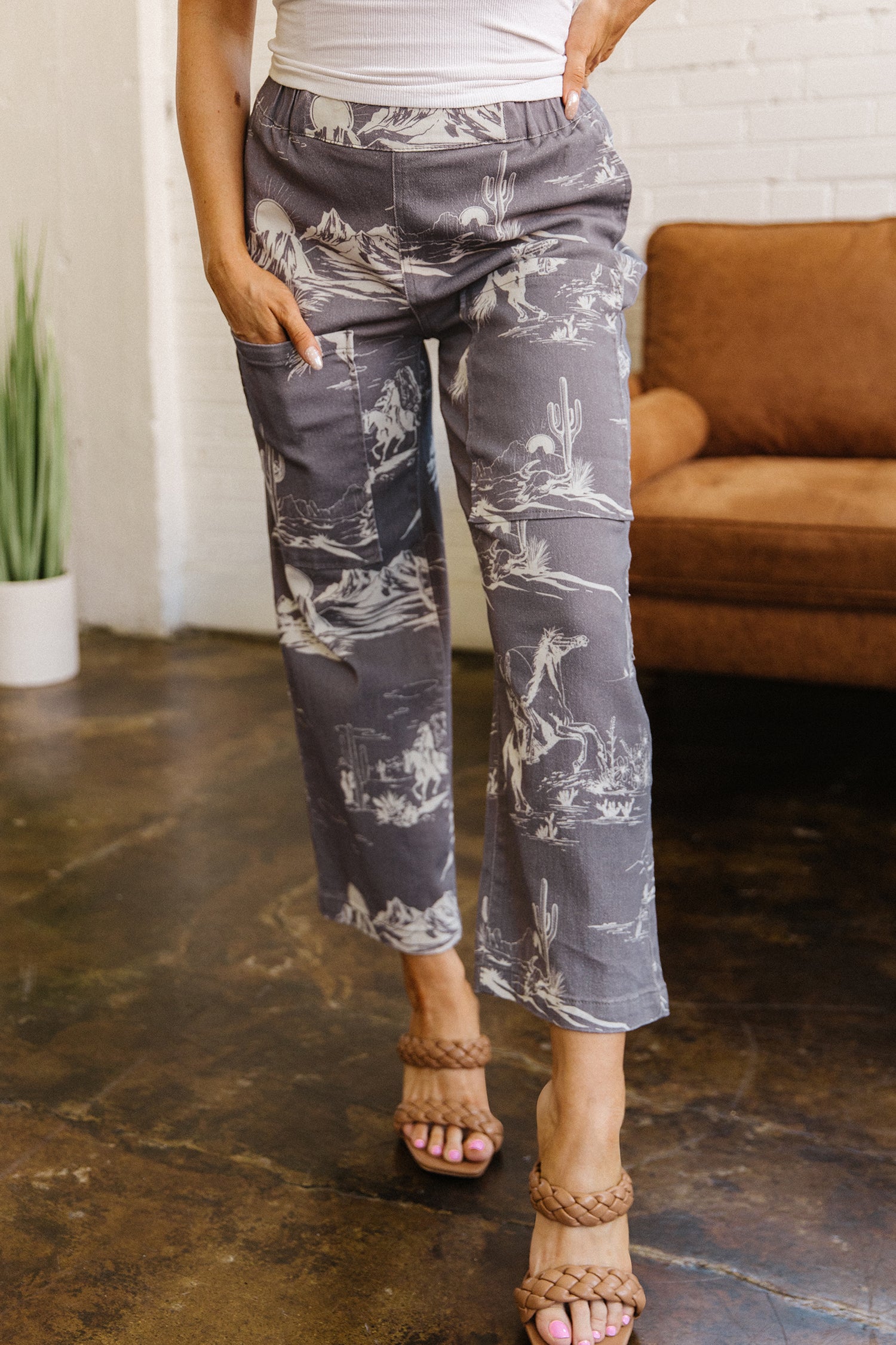 Person wearing gray cowboy print patterned capris in a casual setting with a brown couch and white brick wall.