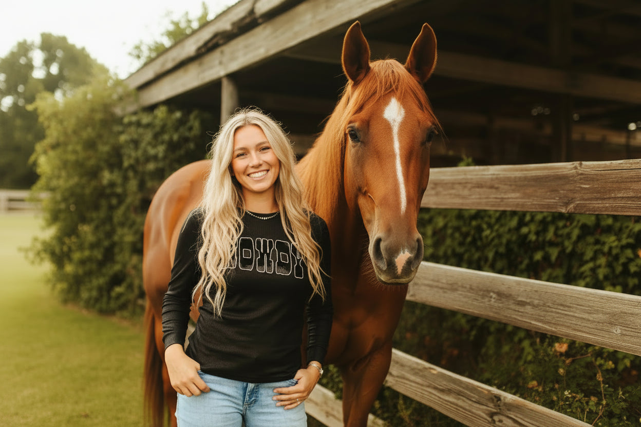 Woman wearing a black long-sleeve shirt with 'HOWDY' printed on it, standing in front of a wooden fence.