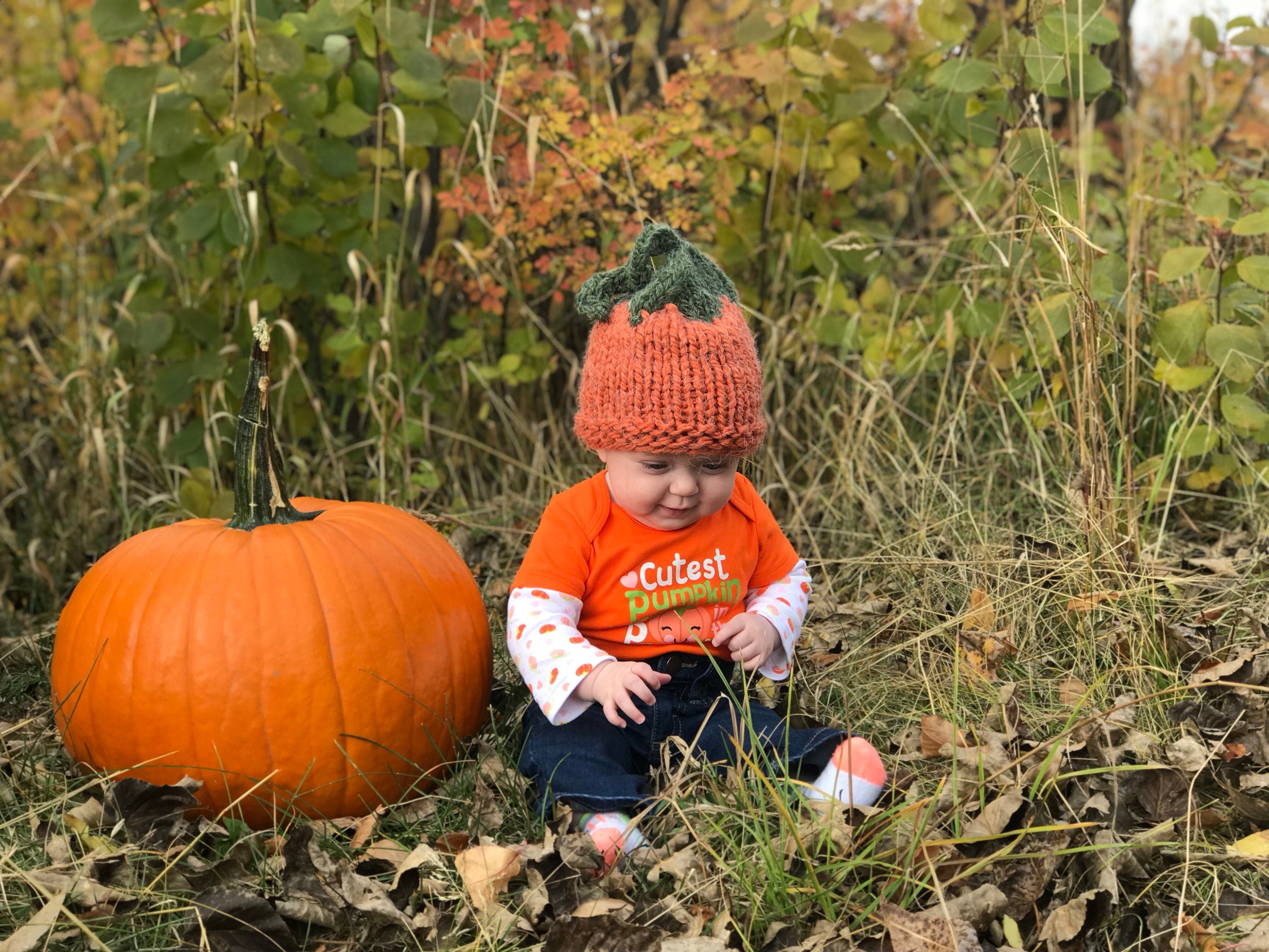 Baby Jernie wearing her handknit pumpkin hat sitting next to a Big pumpkin! Hand-knit pumpkin hat is orange with a green top ending in a pumpkin vine! ๐
