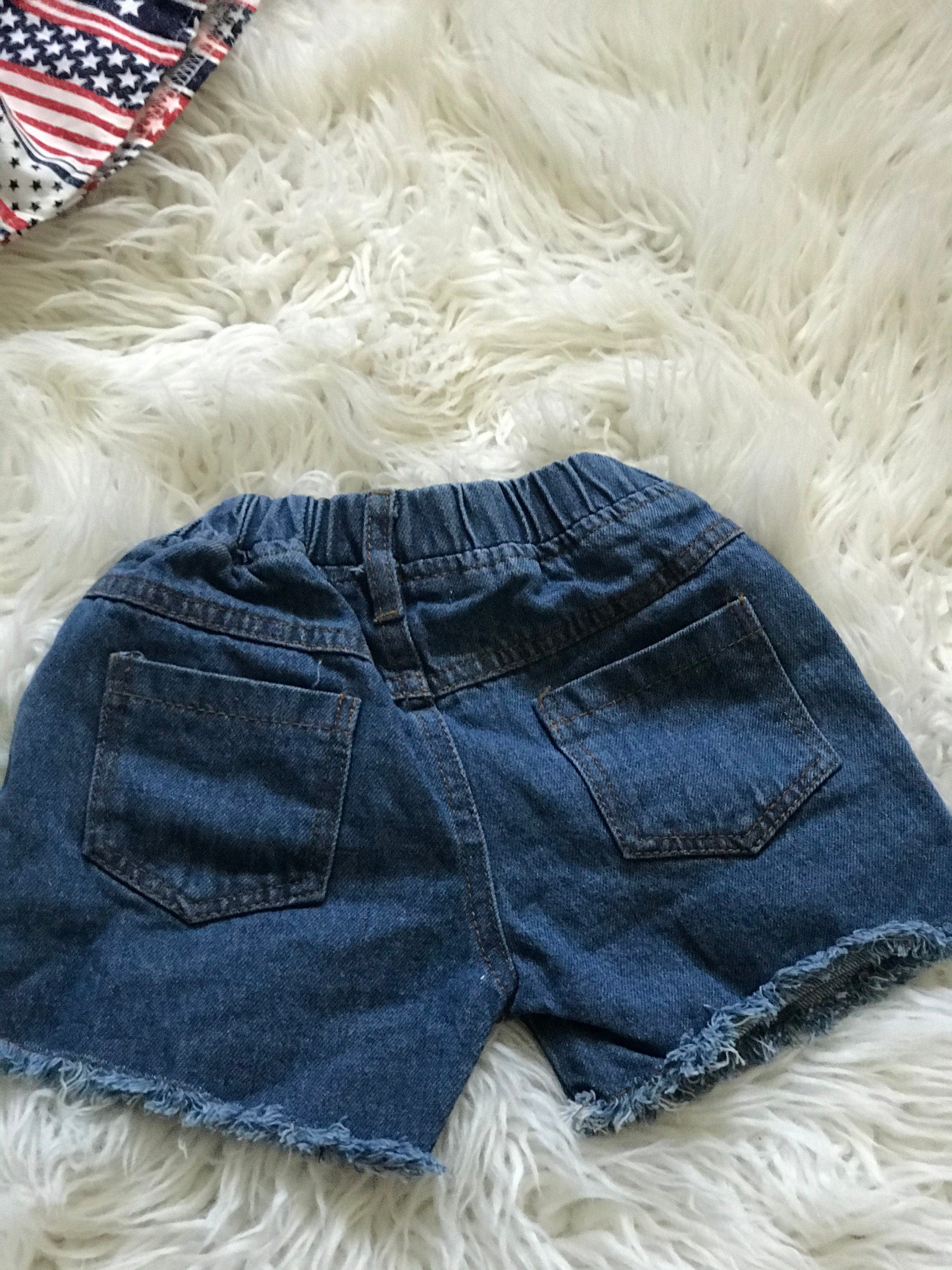 American Girl Short Outfit: The Shorts: The red, white and blue Americana pockets show through the shorts distressing! Shorts have an elastic waist and faux button.
The Shirt: features cold shoulders and an all-around ruffle in USA fabric.