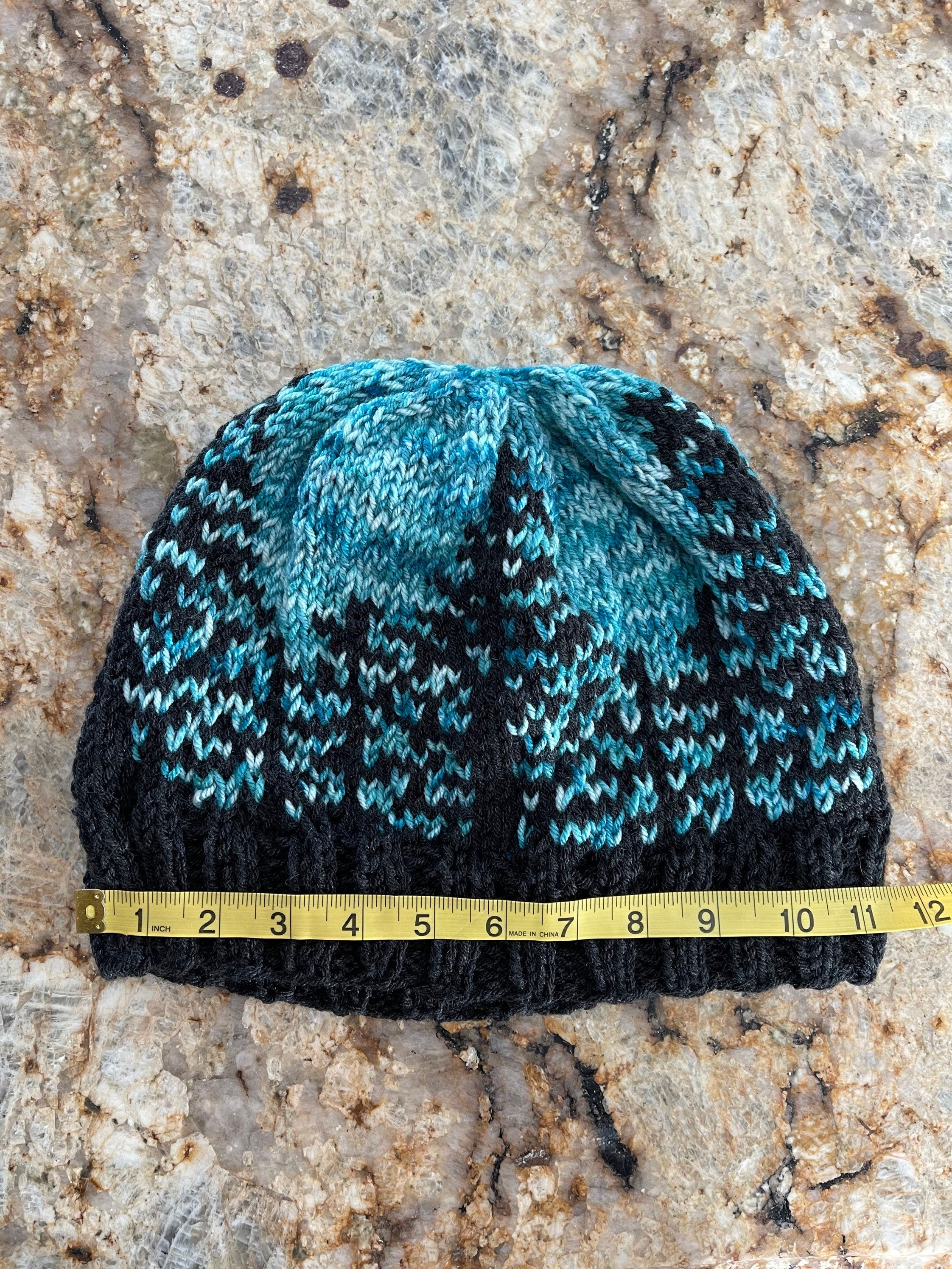Handknit hat featuring a charcoal grey brim and a forest of trees. The background is beautiful turquoise. This hat is ready to ship and a size large.