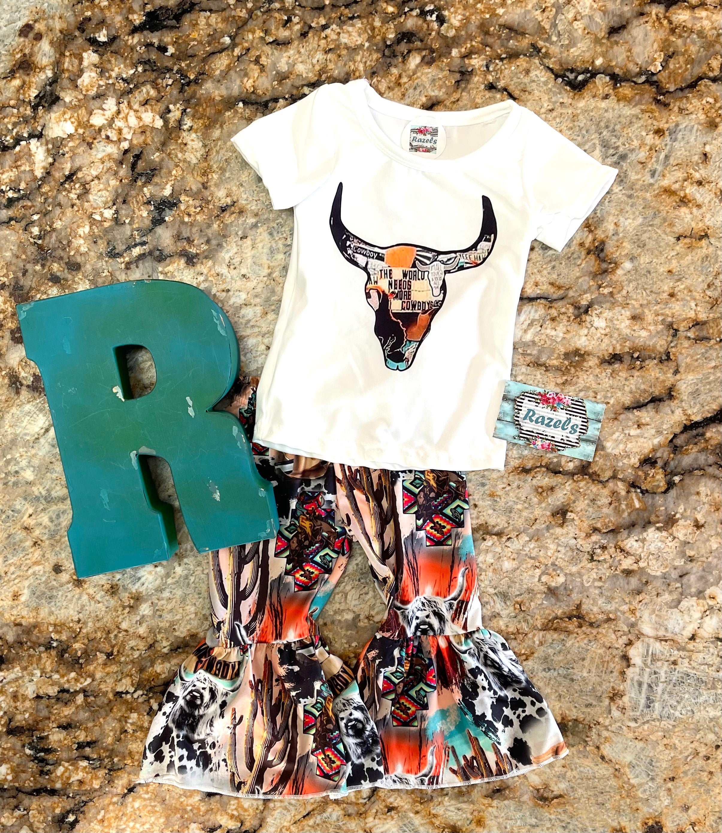 The WorldNeedsMore Cowboys Bell Bottom Outfit, bell bottom pants are a mosaic of Scottish highlander cows, cowprints and cactus. Tshirt features a mosaic inside a cow skull stating the world needs more Cowboys