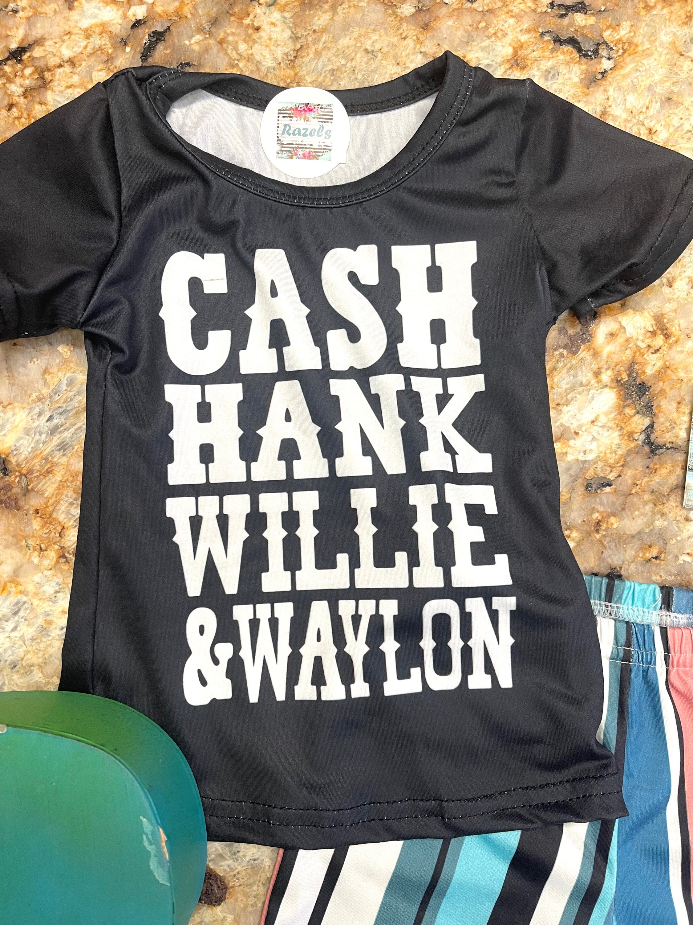 Girls Country Music bell bottom outfit. The outfit features black t-shirt with CASH, HANK, WILLIE, AND WAYLON scrolled across the front. Bell bottom pants feature stripes in pink, blue, black, white and turquoise.