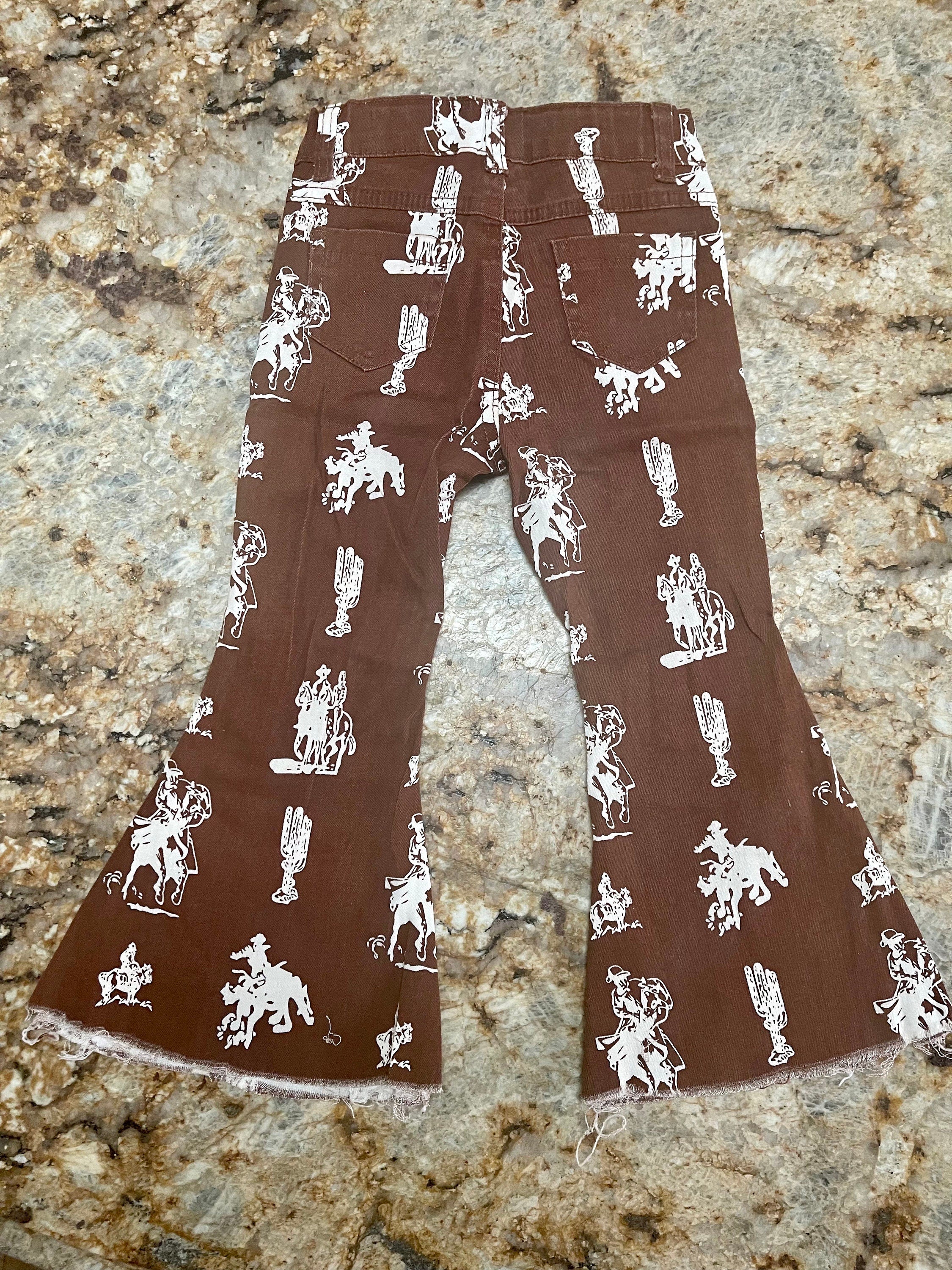 Brown Vintage Cowboy Bell Bottom Jeans featuring a vintage cowboy print, adjustable waistband, functioning pockets, and frayed bells. Styled with a matching Mama's Cowgirl T-shirt. Ideal for dressing up or down, perfect for little cowgirls."