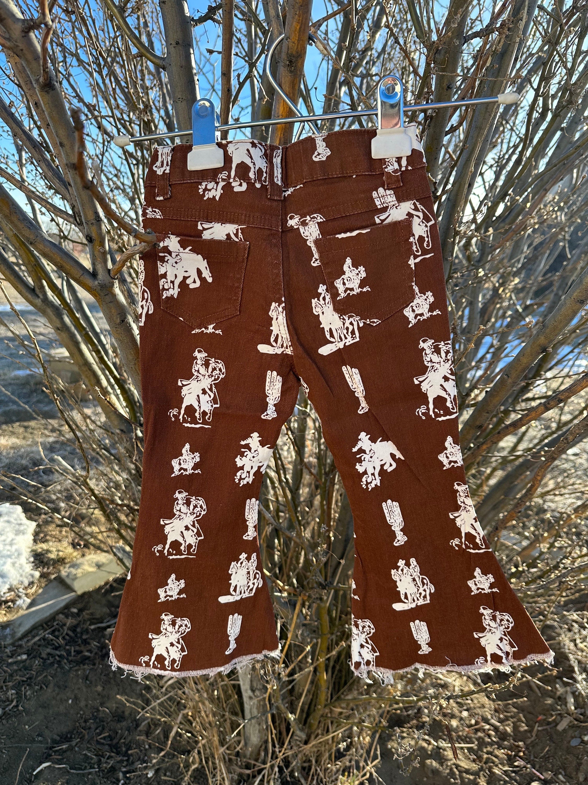 Brown Vintage Cowboy Bell Bottom Jeans featuring a vintage cowboy print, adjustable waistband, functioning pockets, and frayed bells. Styled with a matching Mama's Cowgirl T-shirt. Ideal for dressing up or down, perfect for little cowgirls."