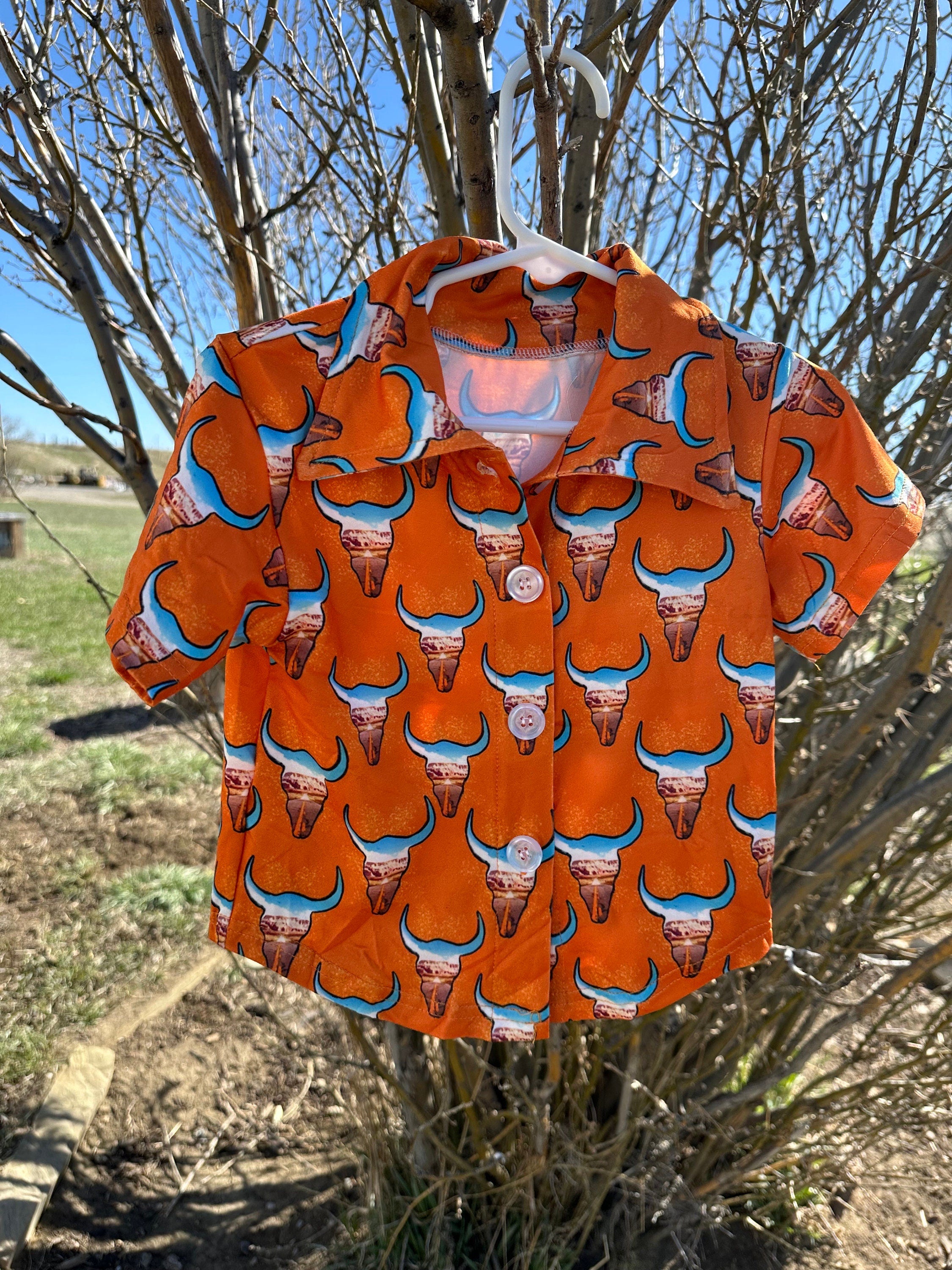 Check out this fun Kids Western button-up shirt featuring a unique desert highway scene inside steer heads. In a bold orange, this cowboy shirt perfectly blends cool and classic Western style—a shirt any cowkid would love! 🧡