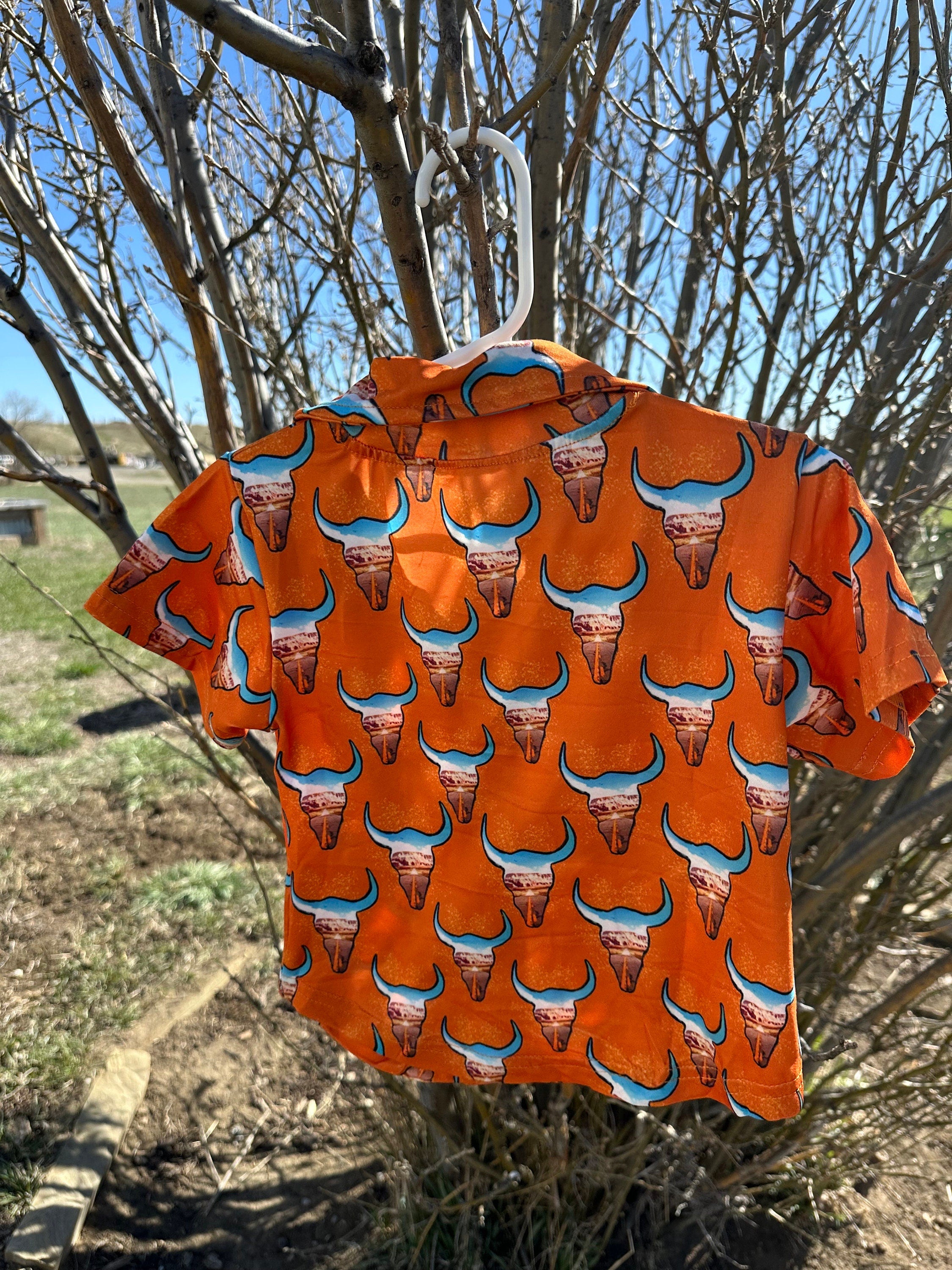 Check out this fun Kids Western button-up shirt featuring a unique desert highway scene inside steer heads. In a bold orange, this cowboy shirt perfectly blends cool and classic Western style—a shirt any cowkid would love! 🧡