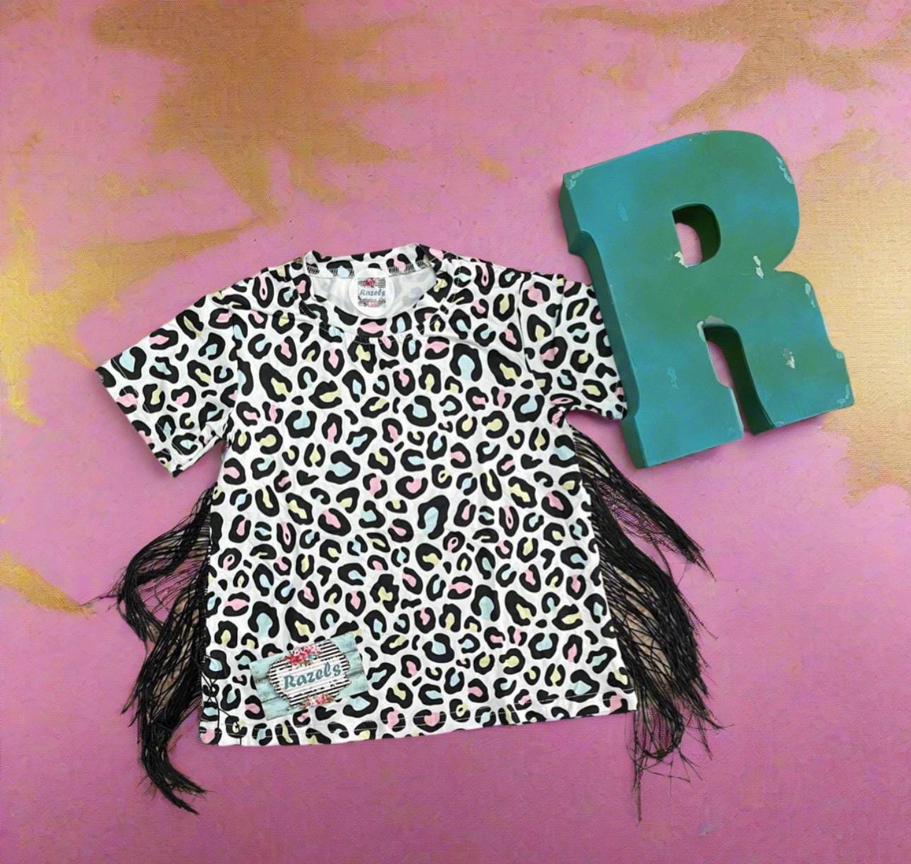 Girls Cheetah Print Fringe T-Shirt Dress: Multicolor cheetah print with pink, yellow, and blue highlighted by black. Adorned with black side fringe down both sides of the dress for a comfy, fun, and adorable look!