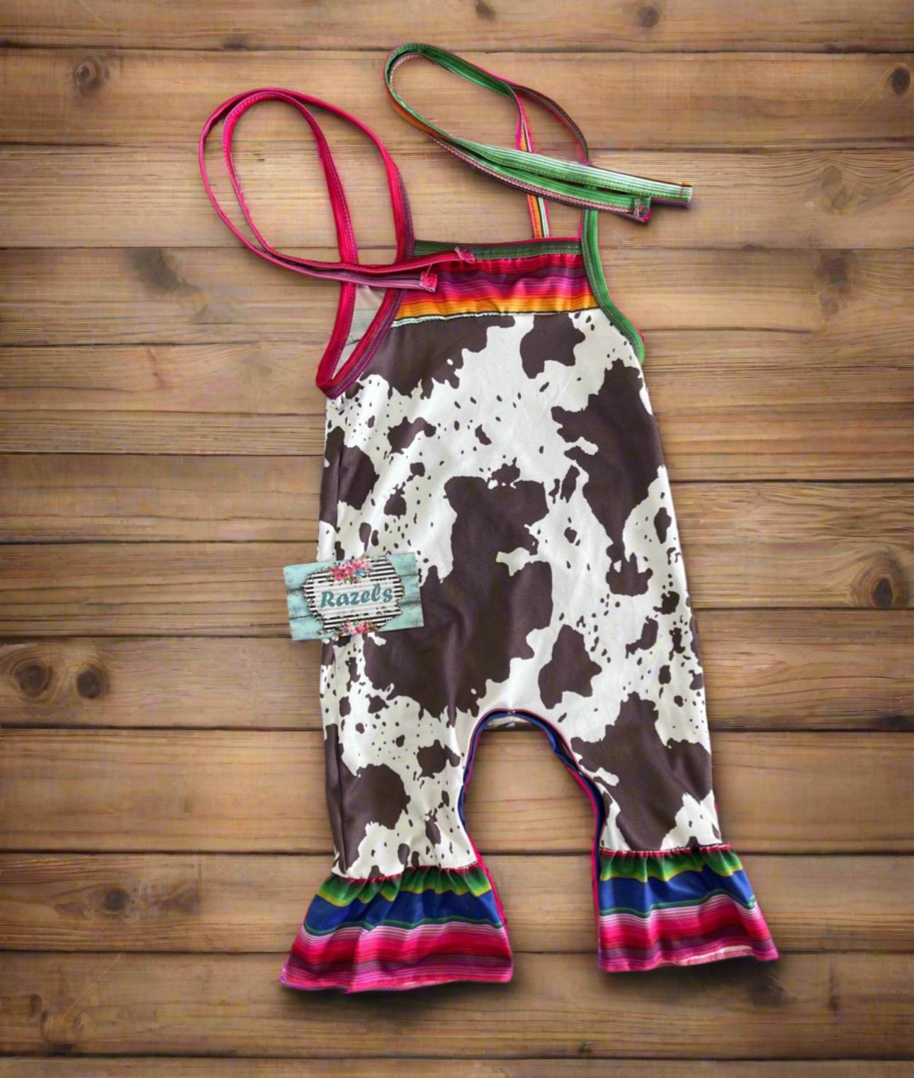 Cowprint Serape Jumpsuit—This stylish Cowprint Outfit features a bold cowprint design complemented by fun pink, blue, and green serape stripes on the neckline, shoulder tie straps, and ruffle hem. Easy 1 piece Cowgirl outfit. Ships FREE with $35+