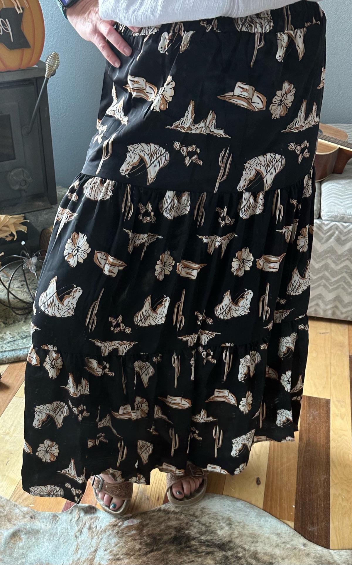 Women's Western BOHO maxi skirt features a charming western print with horses, cacti, flowers, and cowboy hats. It has a high-waisted design and a flowing silhouette in a fall color combo of black, brown, tan, and cream.