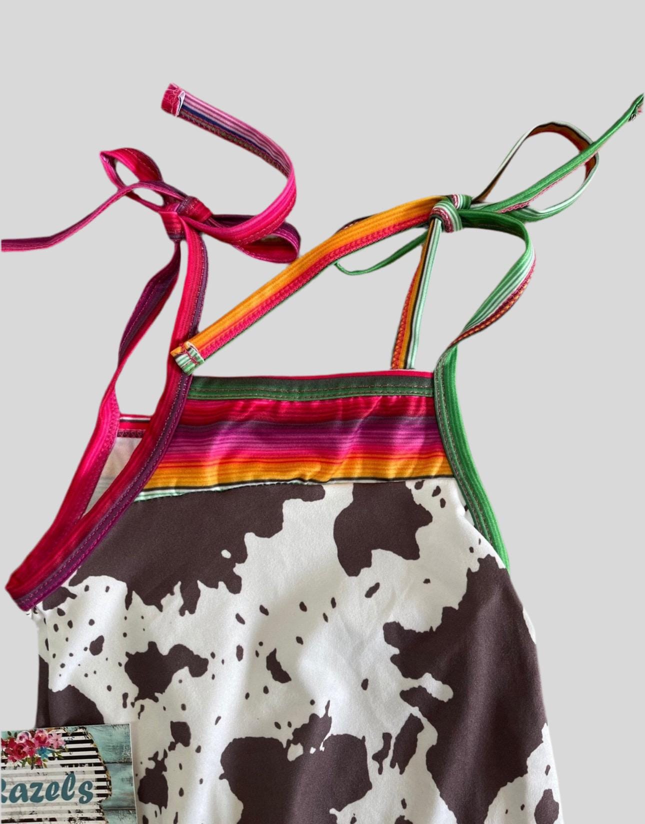 Cowprint Serape Jumpsuit—This stylish Cowprint Outfit features a bold cowprint design complemented by fun pink, blue, and green serape stripes on the neckline, shoulder tie straps, and ruffle hem. Easy 1 piece Cowgirl outfit. Ships FREE with $35+