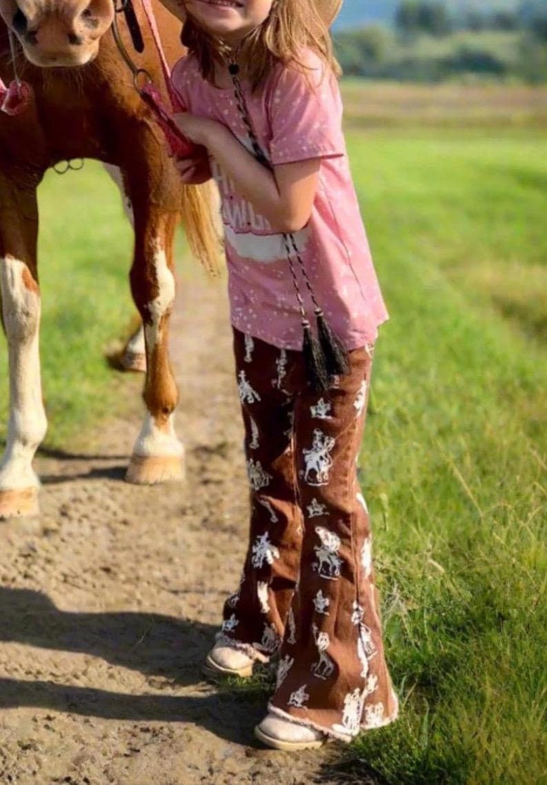 Brown Vintage Cowboy Bell Bottom Jeans featuring a vintage cowboy print, adjustable waistband, functioning pockets, and frayed bells. Styled with a matching Mama's Cowgirl T-shirt. Ideal for dressing up or down, perfect for little cowgirls