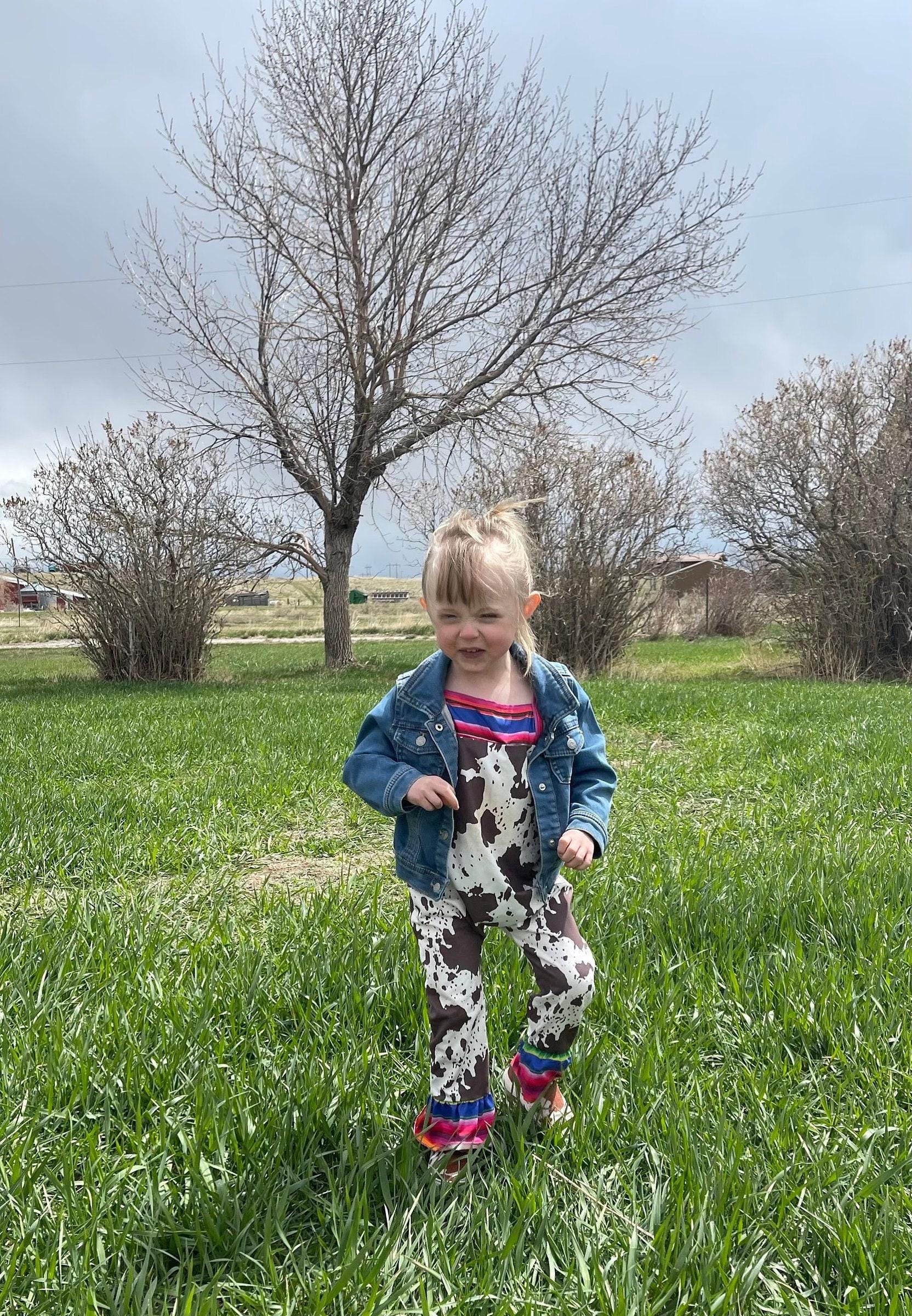 Cowprint Serape Jumpsuit—This stylish Cowprint Outfit features a bold cowprint design complemented by fun pink, blue, and green serape stripes on the neckline, shoulder tie straps, and ruffle hem. Easy 1 piece Cowgirl outfit. Ships FREE with $35+