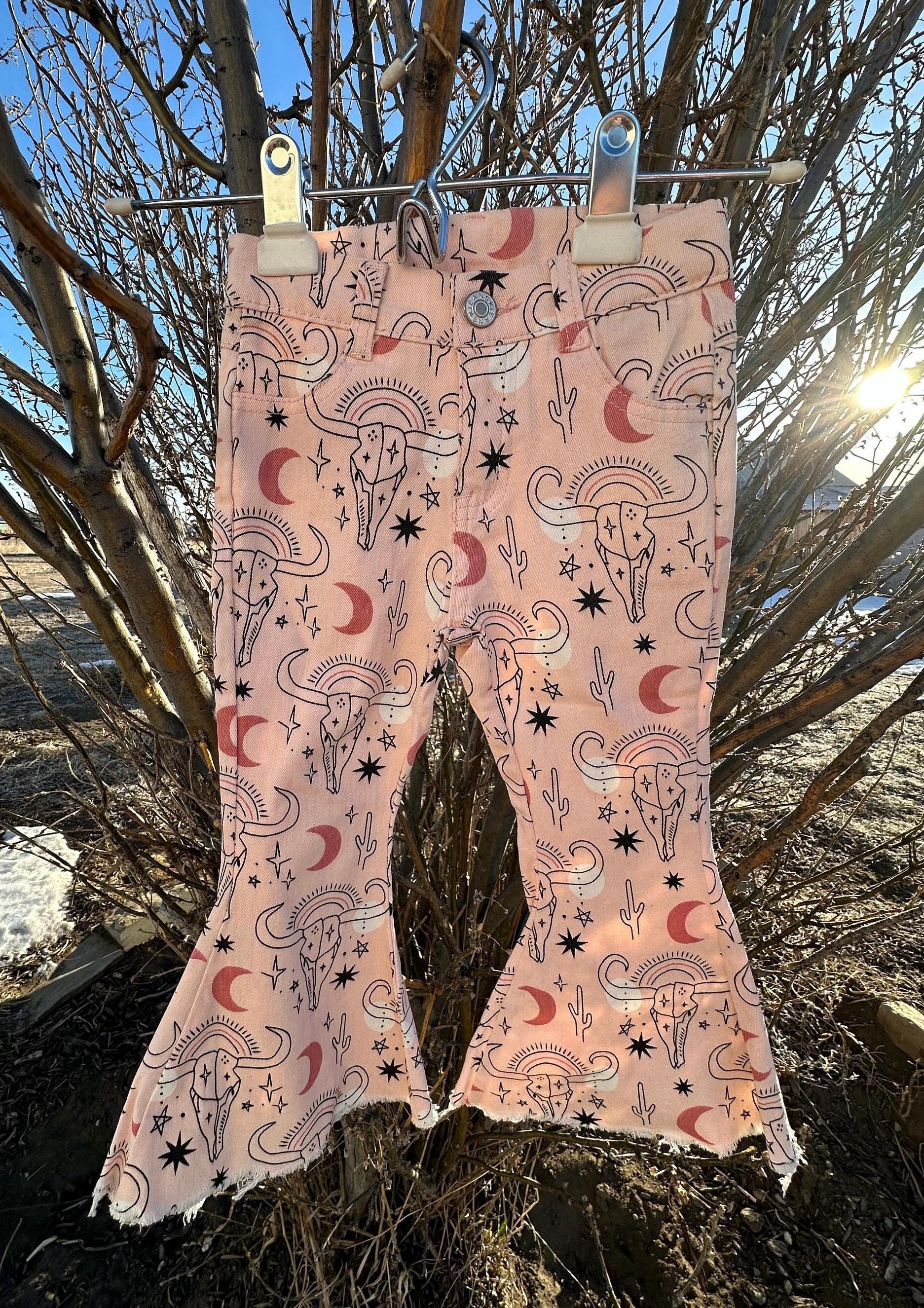 Cowgirl Pink Bell Bottoms – Western Cow Skull Jeans (ONLY 2T, 3T, 4T LEFT!) New cowgirl jeans just dropped—and aren’t they fabulous?! 💓 With cow skulls, moons, cactus, stars... oh my! These pink bell bottoms are full of western sass and sweet desert vibes. ✓ Functional front and back pockets ✓ Adjustable waistband for a perfect fit ✓ Stretchy cotton/spandex blend for all-day comfort ✓ Button and zipper closure ✓ Frayed hems for that trendy flair