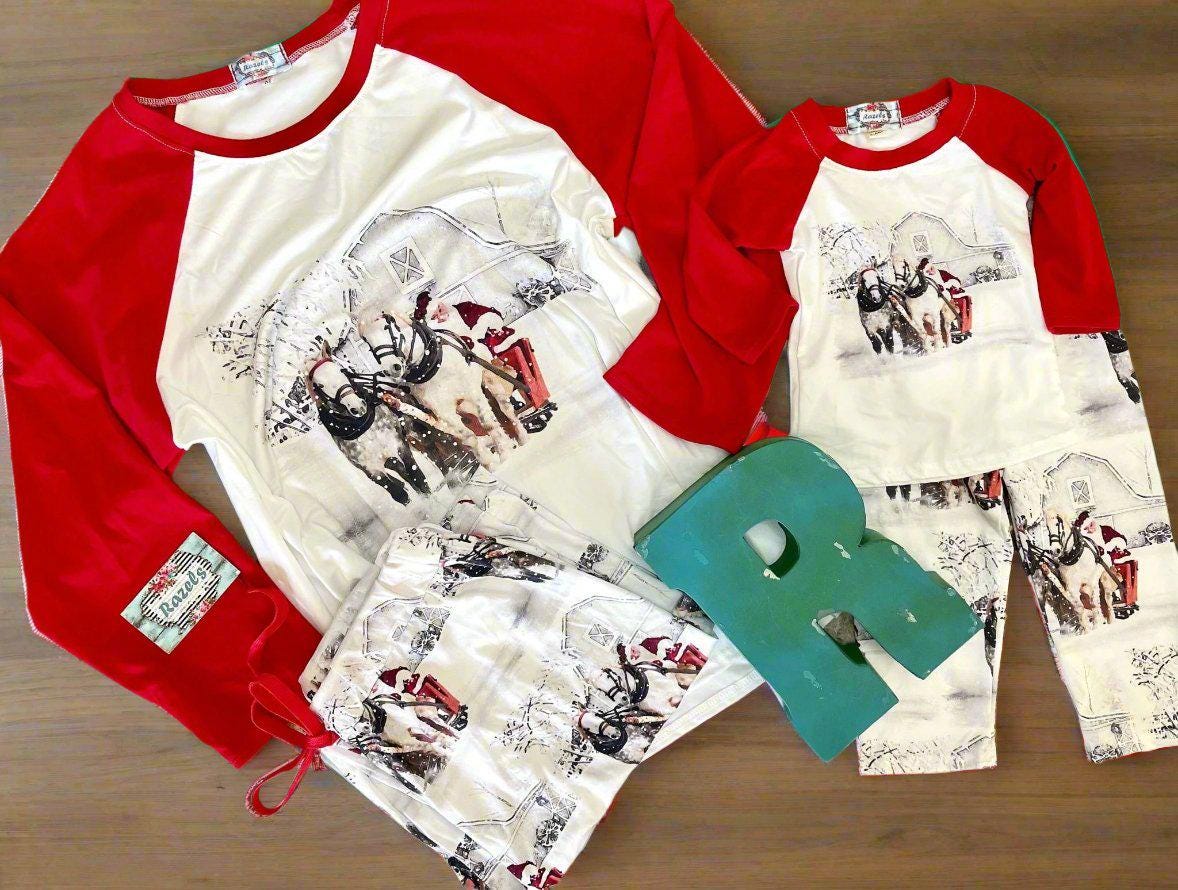 Family White Christmas Pajamas feature Santa Claus at the reins of a horse-drawn sleigh, pulling away from a vintage barn on a snowy winter day. The top has coordinating red raglan sleeves, and the bottoms feature a festive Santa print all over