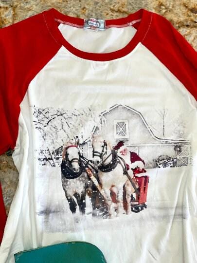 Family white Christmas Pajamas feature Santa Claus at the reins of a horse-drawn sleigh, pulling away from a vintage barn on a snowy winter day. The top has coordinating red raglan sleeves, and the bottoms feature a festive Santa print all over