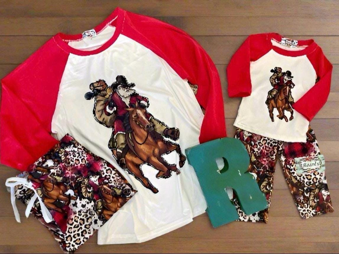 family cowboy Santa Christmas pajamas feature a raglan-style top with Santa riding his best horse, carrying a sack of packages. The top has red sleeves and neckline. The bottoms have an all-over cheetah print, buffalo plaid and Santa on his horse
