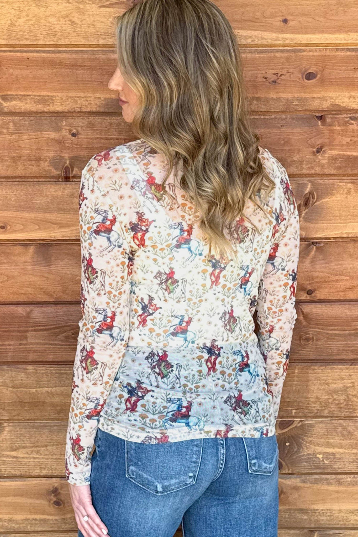 A woman viewed from the back wearing a long-sleeve cream-colored blouse with a colorful mesh cowboy and floral print, paired with blue jeans, standing against a wooden plank wall.