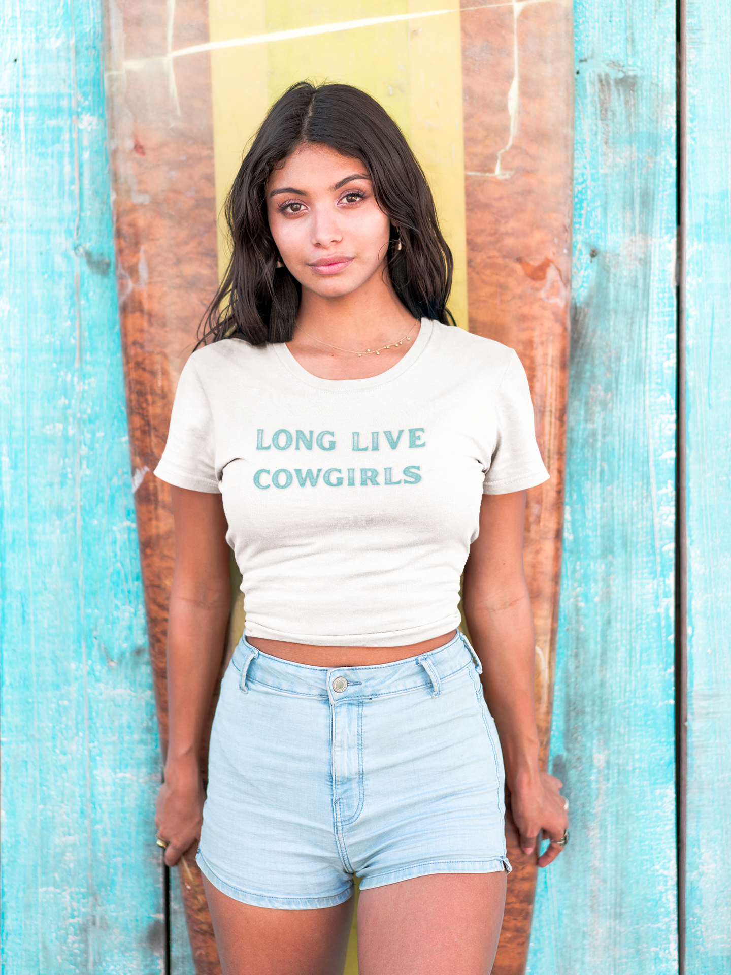 A woman wearing a beige crop top with 'LONG LIVE COWGIRLS' printed in turquoise letters on the front, paired with blue denim shorts.