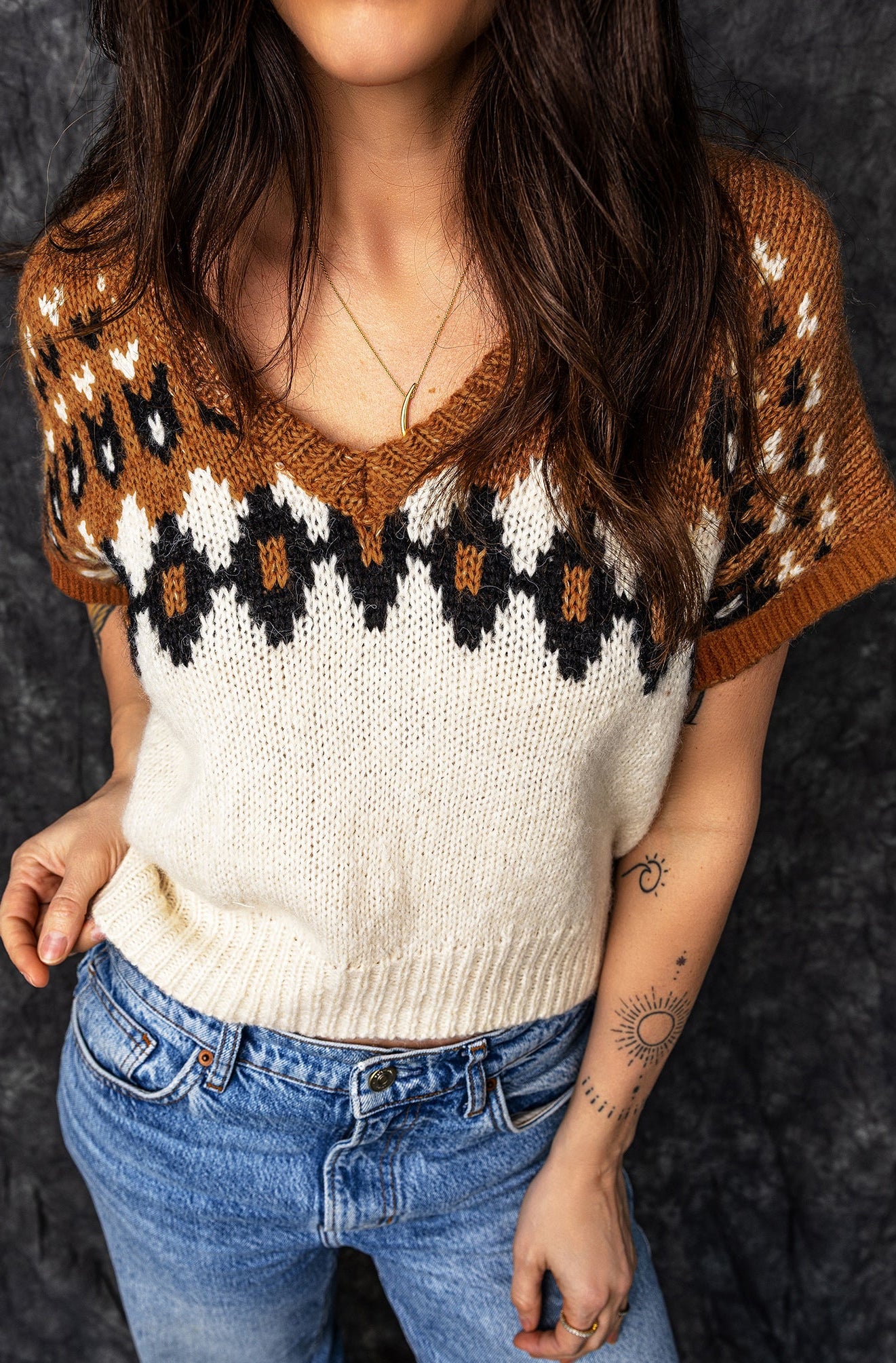 A woman wearing a short-sleeve knit sweater with a brown, cream, and black geometric pattern, paired with blue jeans, standing against a dark textured background.