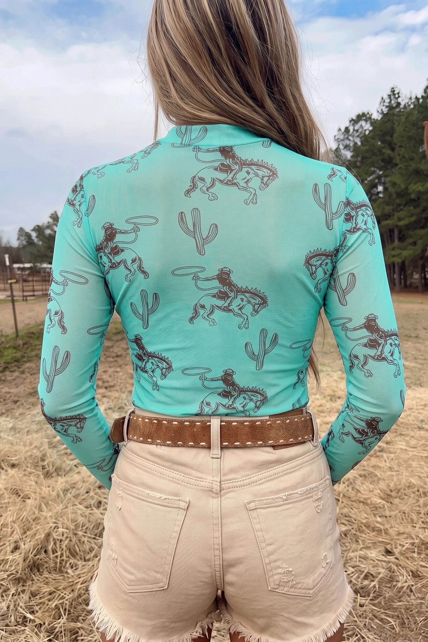 A woman viewed from behind wearing a turquoise long-sleeve shirt with a brown desert cowboy and cactus pattern, paired with beige shorts and a brown belt, standing outdoors in a field.