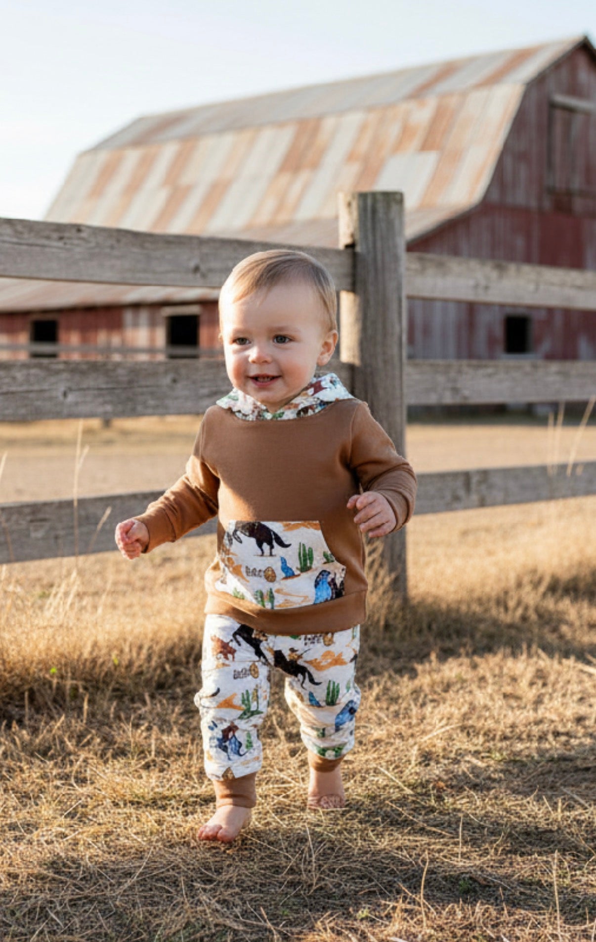 Baby western cowboy jogger pants and hoodie standing in a field with a barn in the background