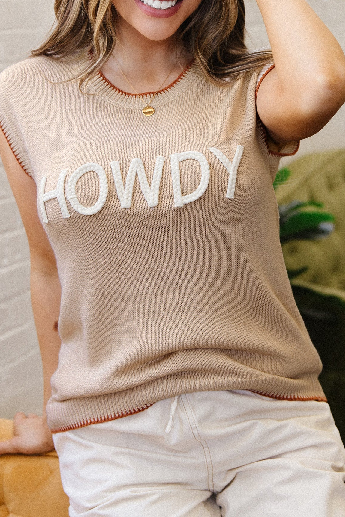 Woman wearing a beige sleeveless sweater with 'HOWDY' text, sitting on a yellow couch.