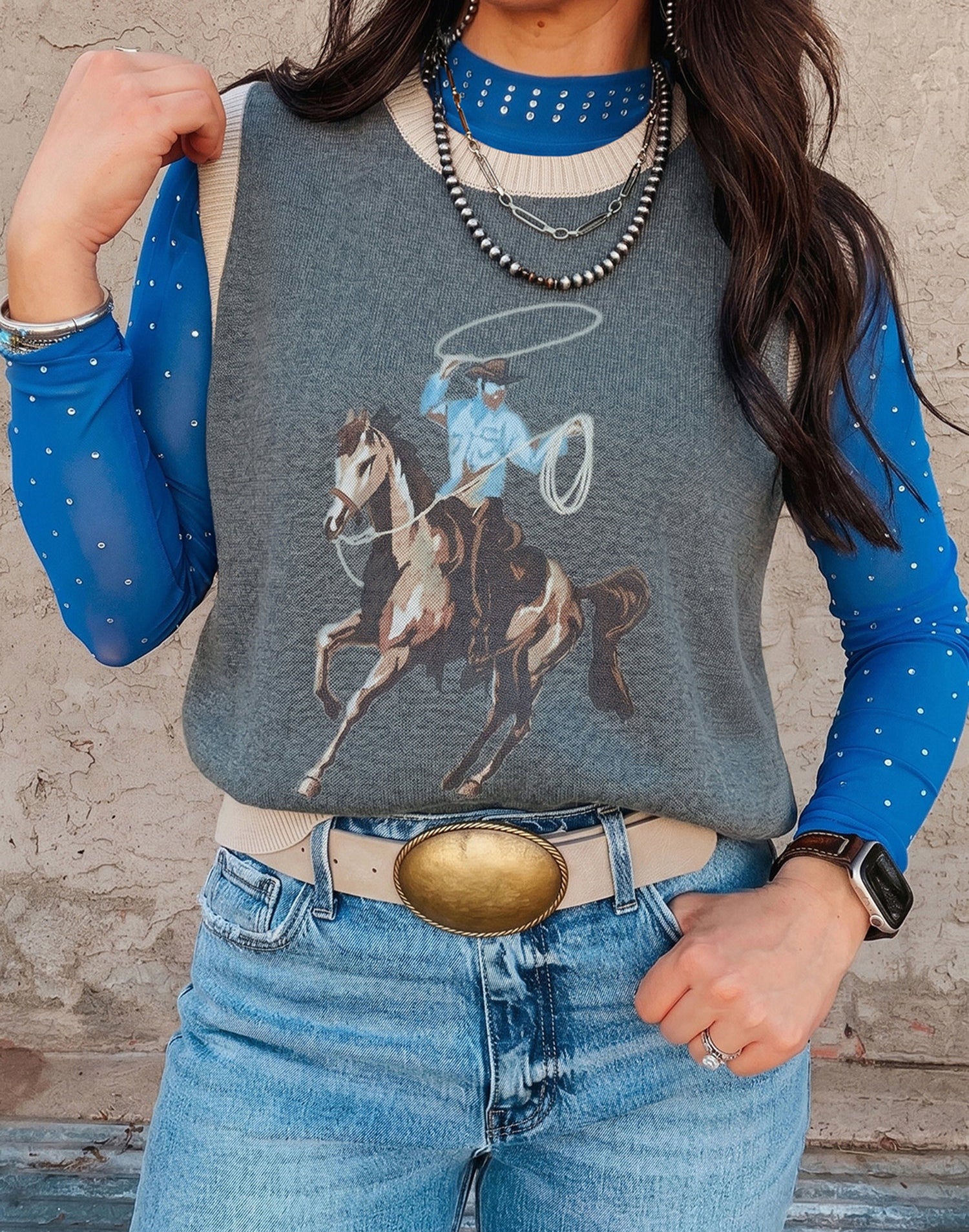 women wearing a gray long-sleeve shirt with a horse and lasso graphic, blue jeans, and a belt with a large buckle.