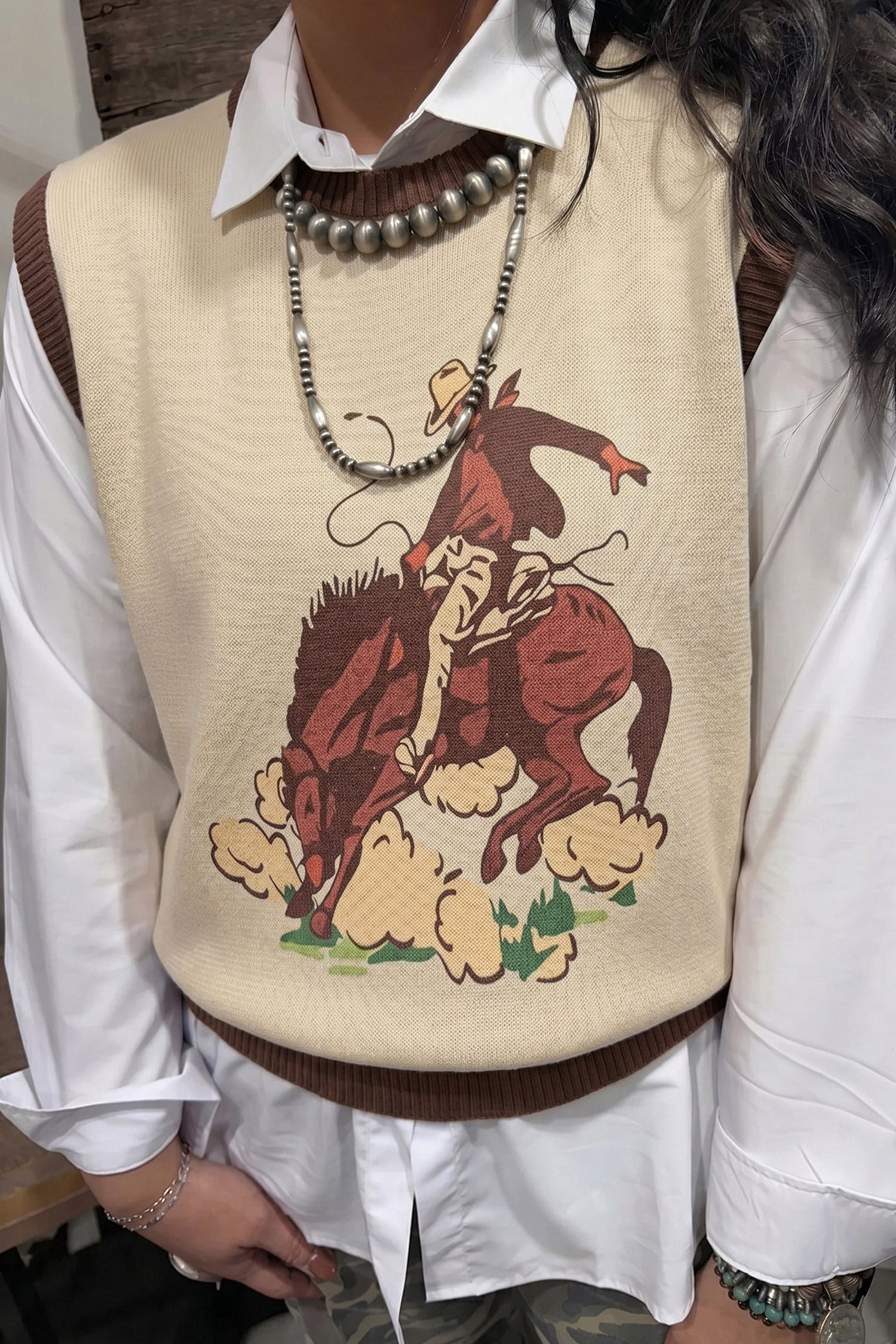closeup of womens western vest featuring a bronc riding cowboy on beige background and brown hem and neck accents. Great alone or layered this fall.