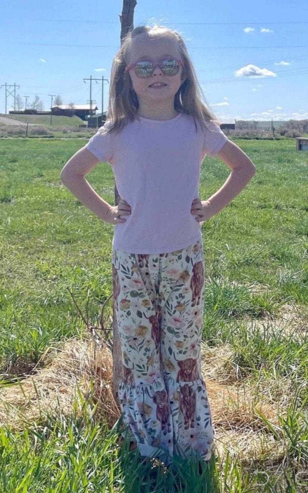 GIRL LEANING AGAINST TREE WEARING A PINK TSHIRT, PINK SUNGLASSES AND Highland Cow Floral Bell Bottoms – Adorable Western Cow Pants for Little GIRLS