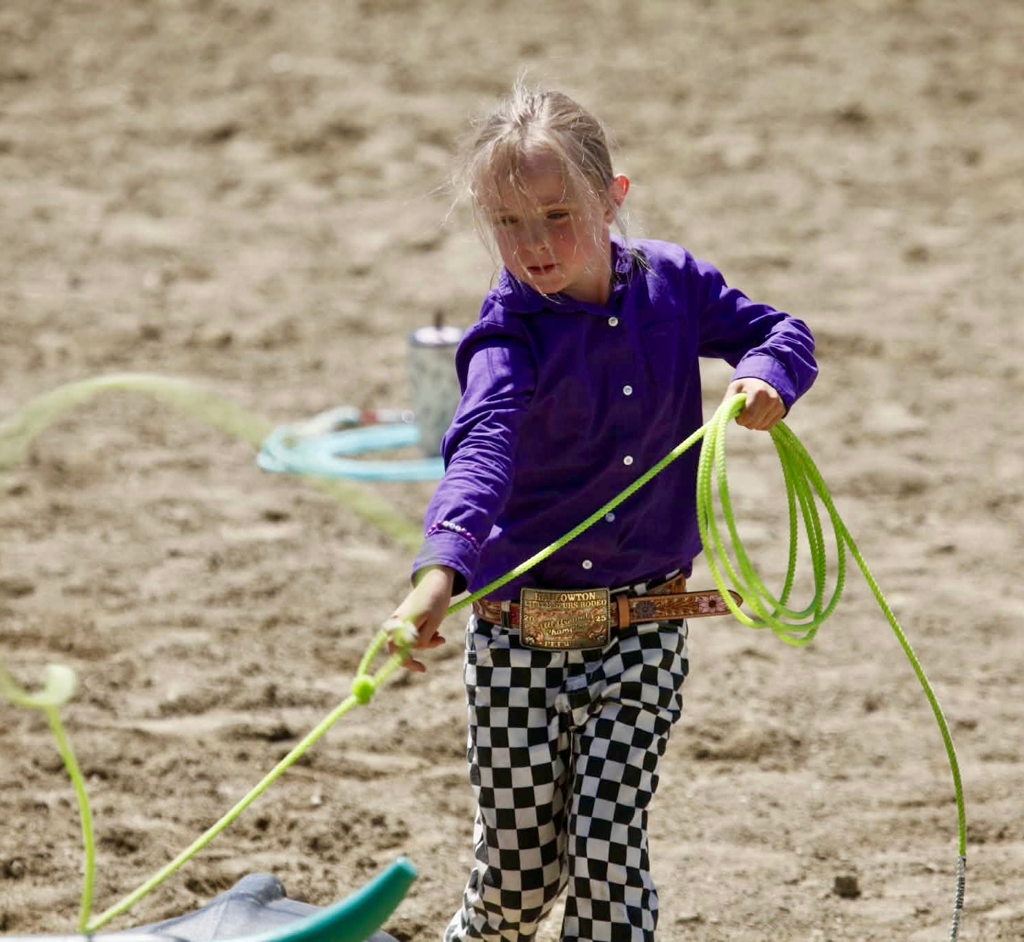 A young girl wearing a purple button-up shirt and black-and-white checkered pants holds a bright green lasso rope on a sandy outdoor ground.