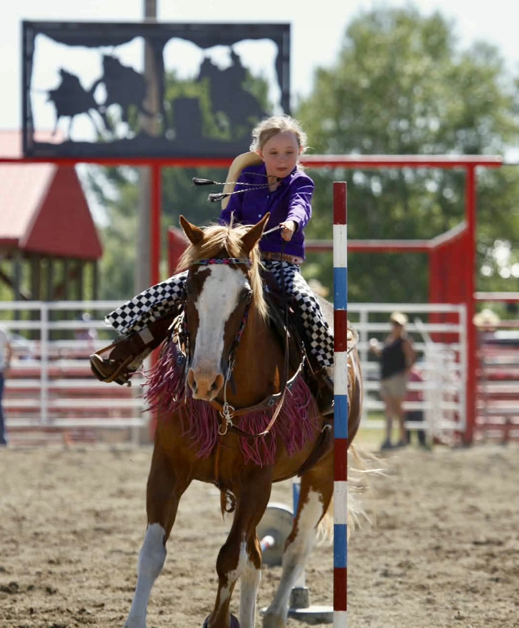 A young girl in a purple shirt and black-and-white checkered pants rides a brown and white horse during an equestrian event in an arena, with a red gate and trees in the background.