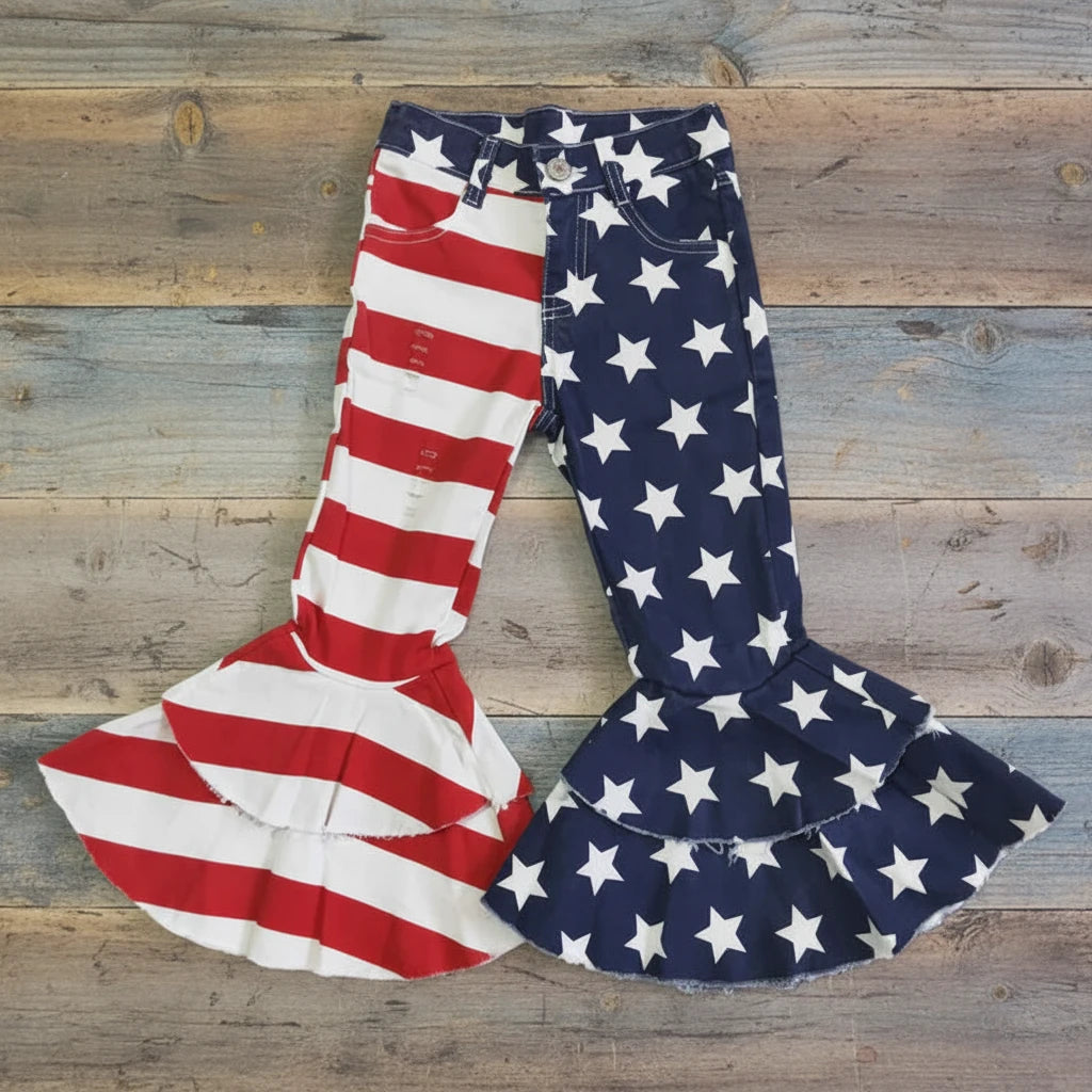 Girls American flag bell bottom jeans featuring a split stars-and-stripes design with one leg in navy star print and the other in red and white stripes. Made from stretch denim with a mid-rise fit, functional front and back pockets, and belt loops. Adjustable waistband with button and zipper closure for a better fit. Finished with a tiered ruffle bell bottom hem for added flare. Available in sizes 6–12M through 14/16.