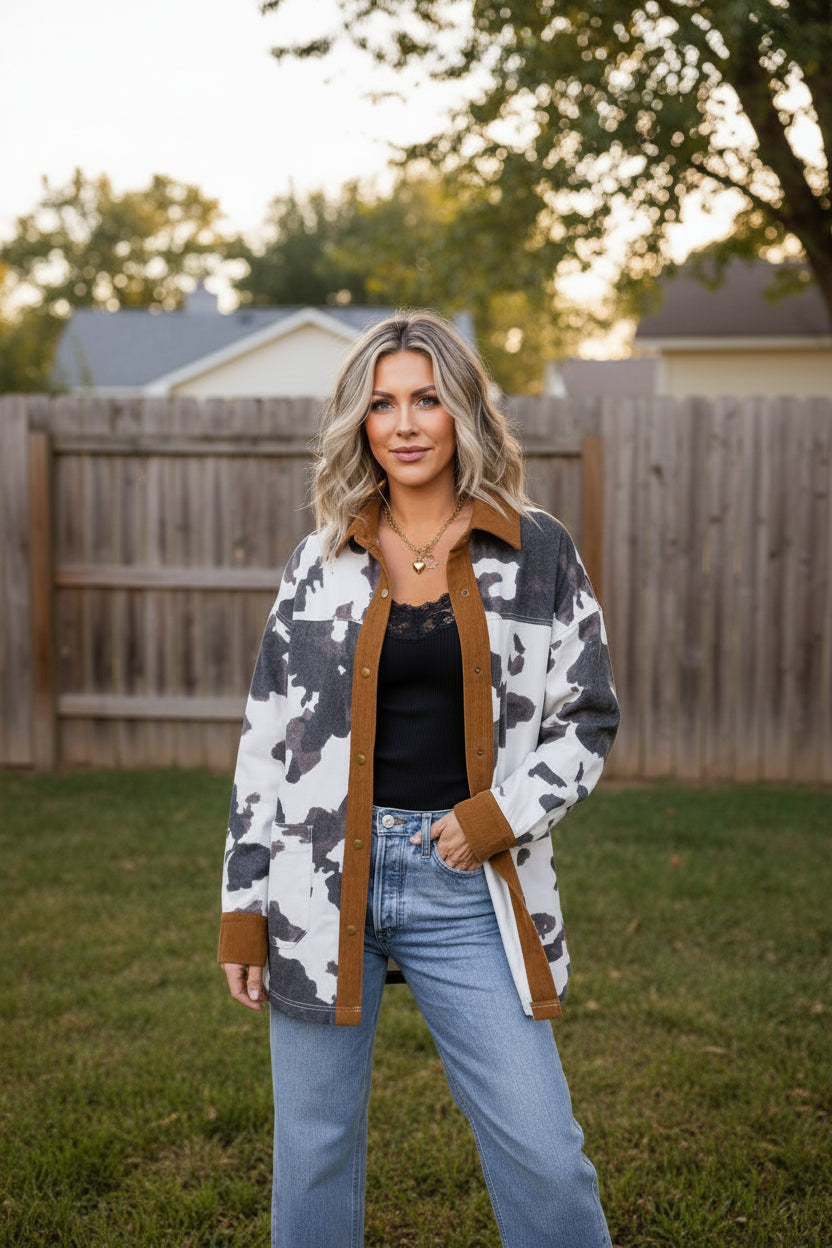 A woman wearing a long-sleeve button-up shirt jacket with a brown and black cow print pattern, brown suede-like collar, cuffs, and front placket, styled over a black top and blue jeans standing outdoors in the fall
