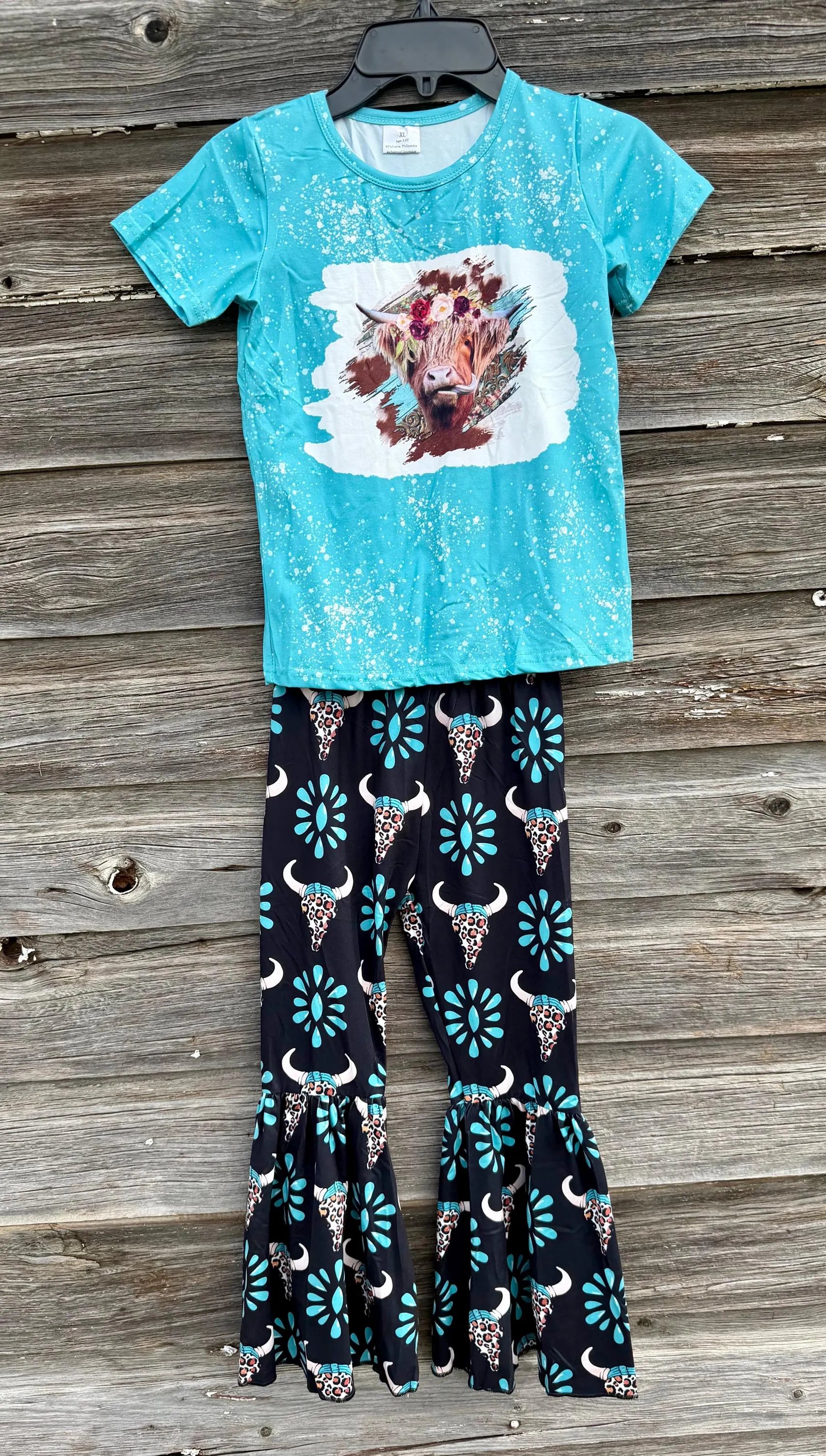 A two-piece girls' outfit set featuring a turquoise short-sleeve t-shirt with a bull graphic and white speckled pattern, paired with black flared pants decorated with turquoise, white, and leopard print details, displayed on a wooden background.