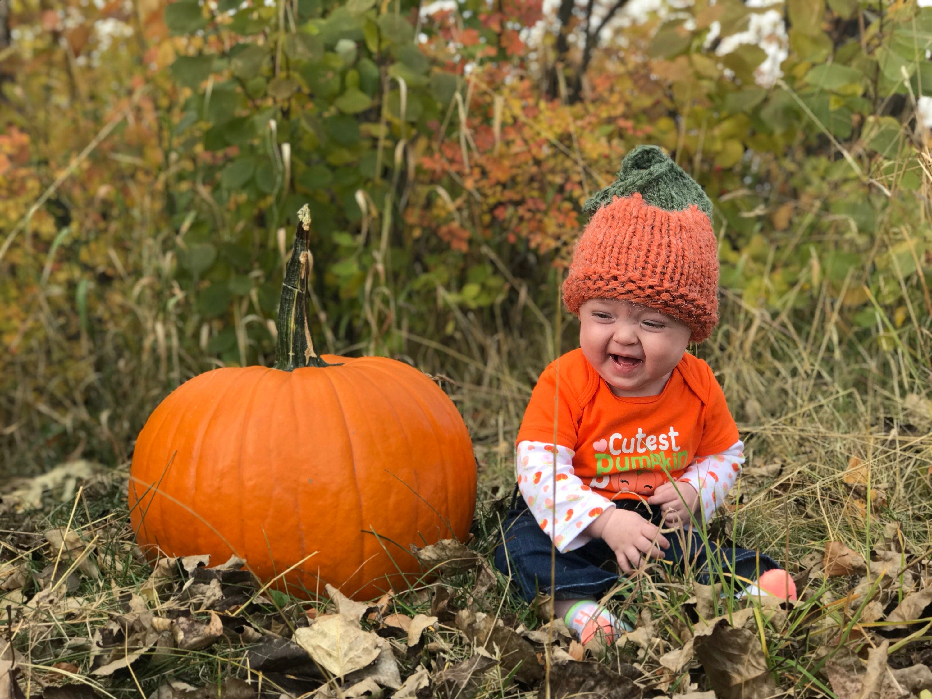 Baby Jernie wearing her handknit pumpkin hat sitting next to a Big pumpkin! Hand-knit pumpkin hat is orange with a green top ending in a pumpkin vine! 🎃