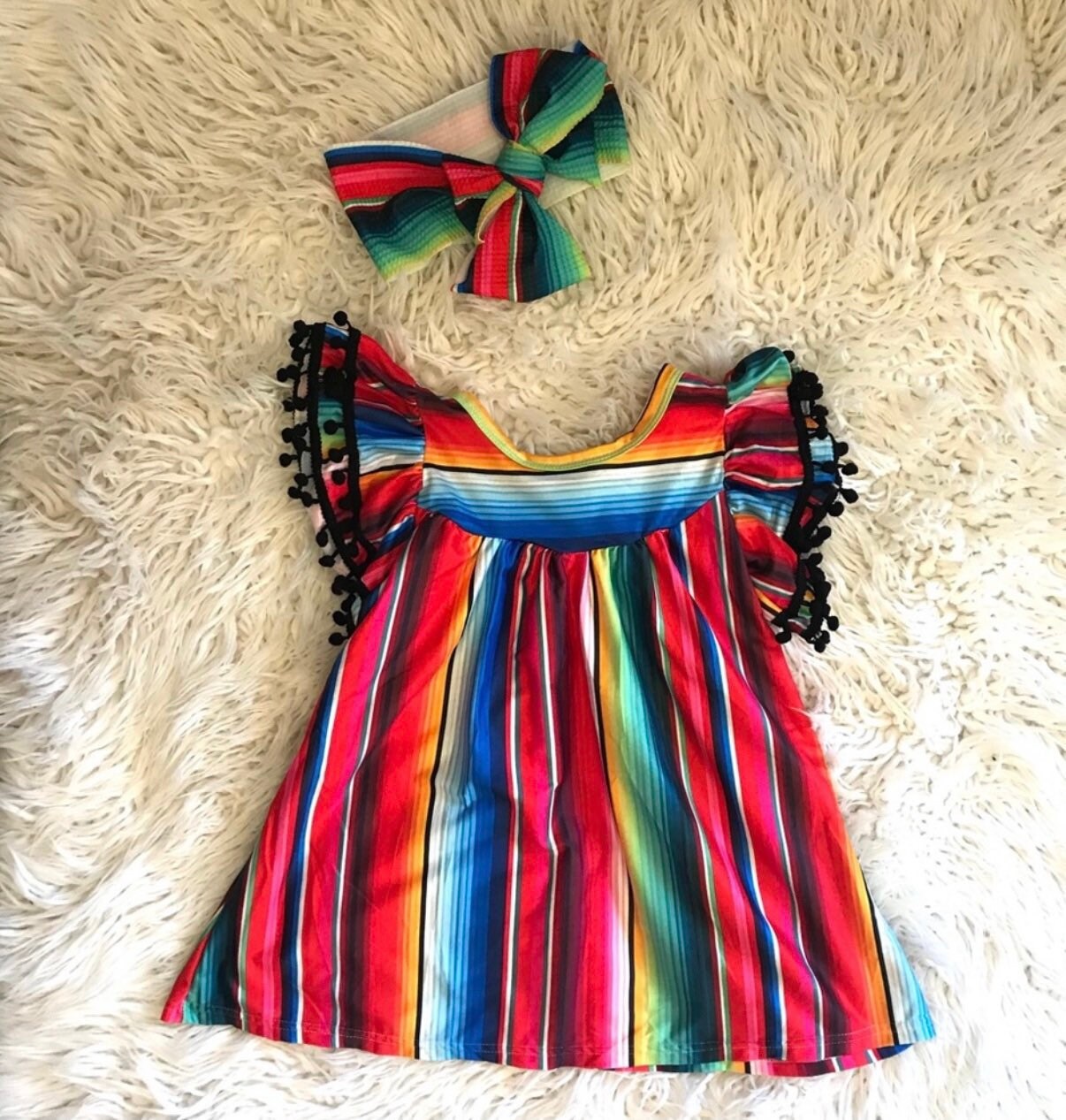 The girls&#39; fiesta party dress features a bright serape stripe throughout and pom-pom cap sleeves.Matching bows are also available