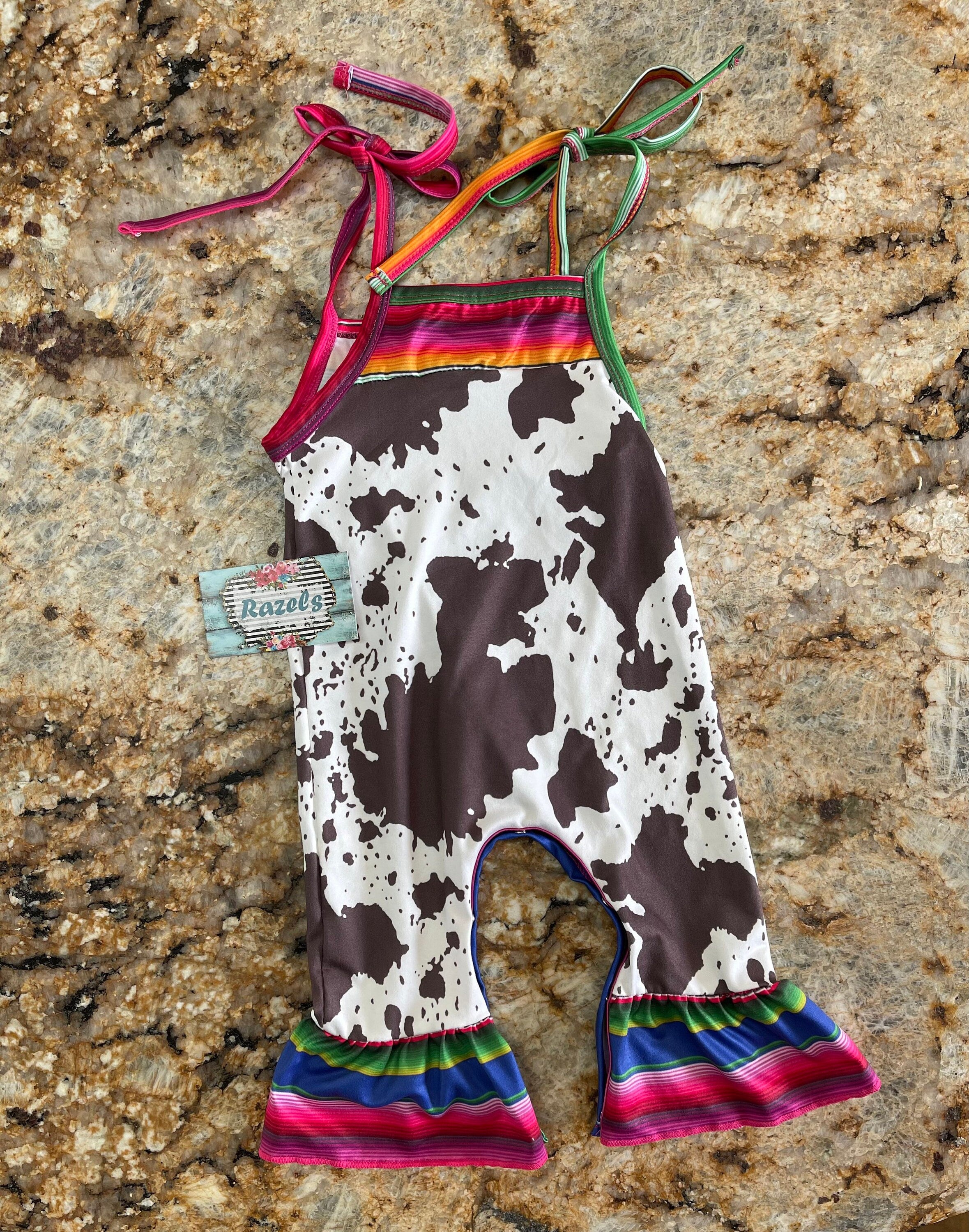 Cowprint Serape Jumpsuit—This stylish Cowprint Outfit features a bold cowprint design complemented by fun pink, blue, and green serape stripes on the neckline, shoulder tie straps, and ruffle hem. Easy 1 piece Cowgirl outfit. Ships FREE with $35+